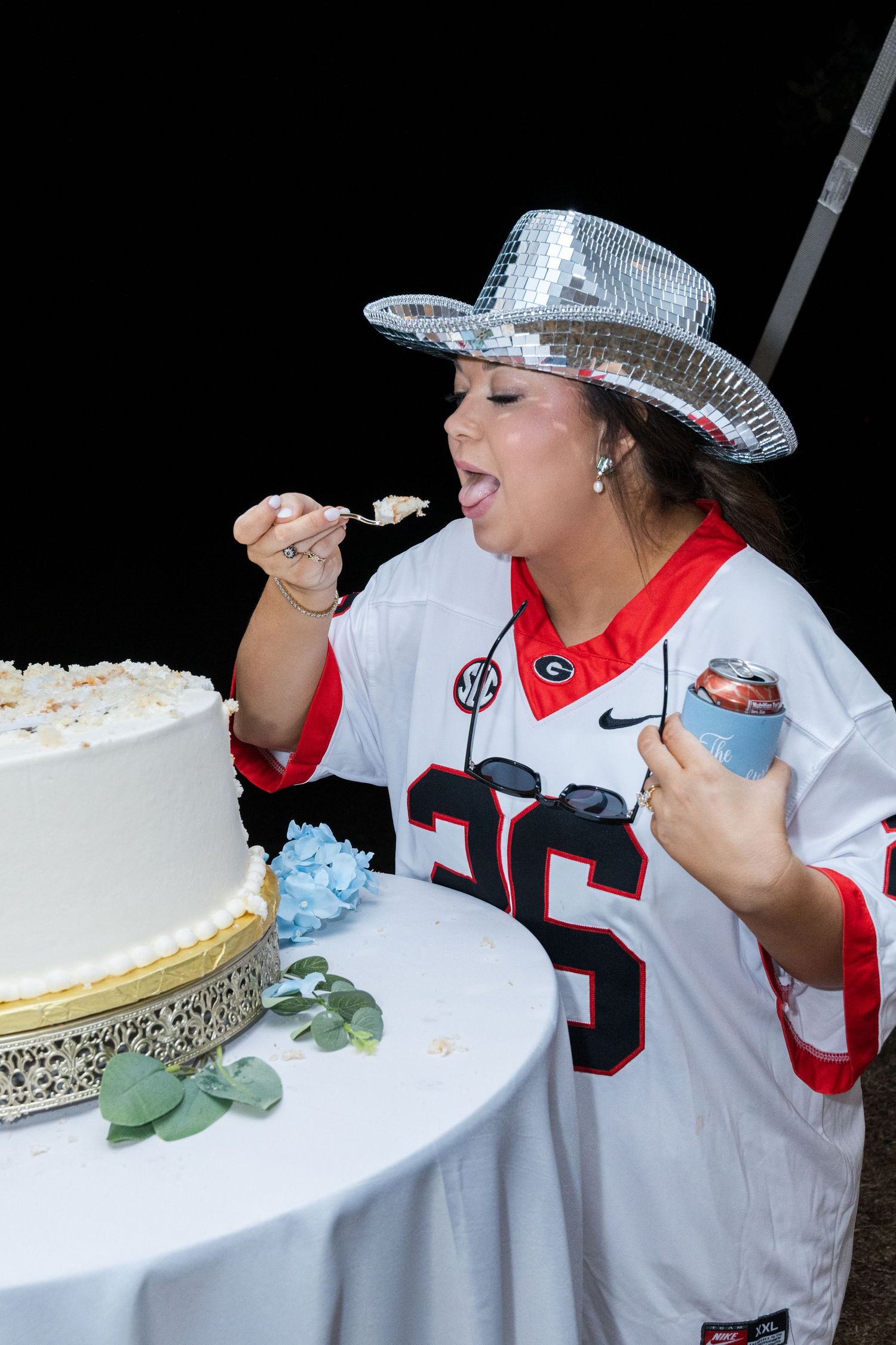 Woman in Georgia Bulldogs jersey and silver hat tastes wedding cake with a spoon at a celebration event.