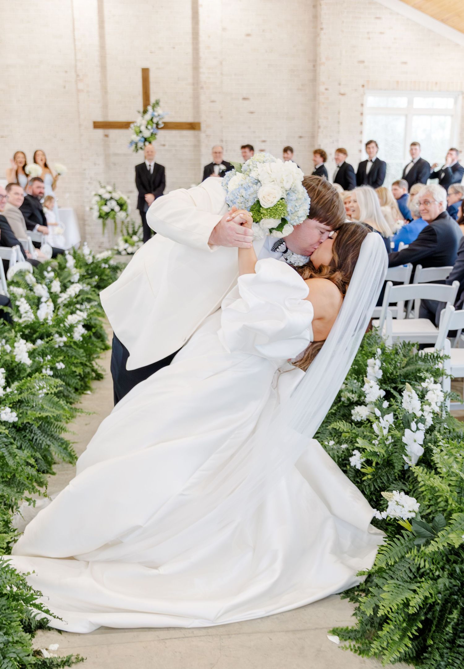 Bride and groom share first kiss at altar in white brick chapel with greenery-lined aisle.