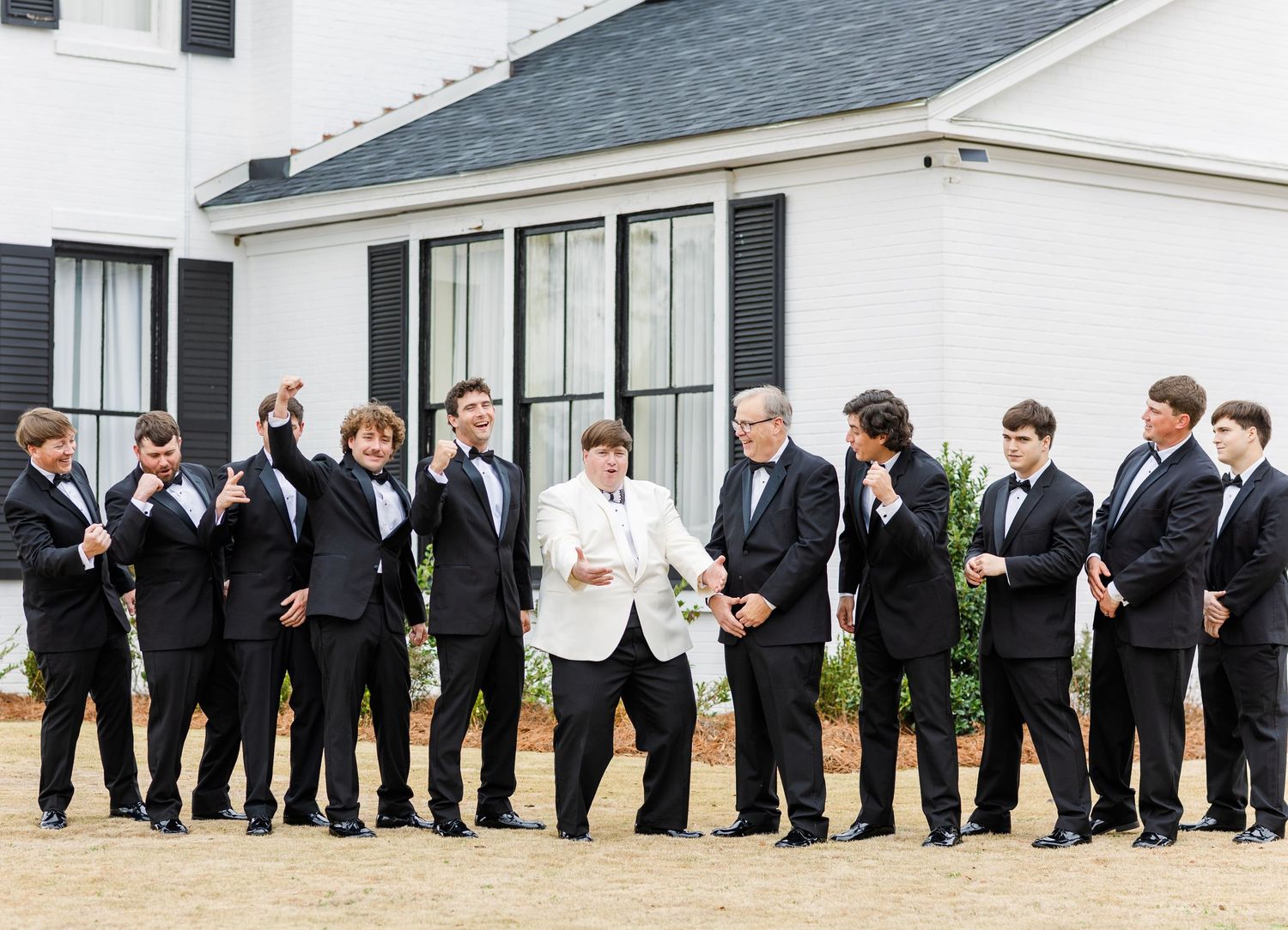 Groom in white jacket celebrates with groomsmen in black tuxedos outside a modern white venue building.