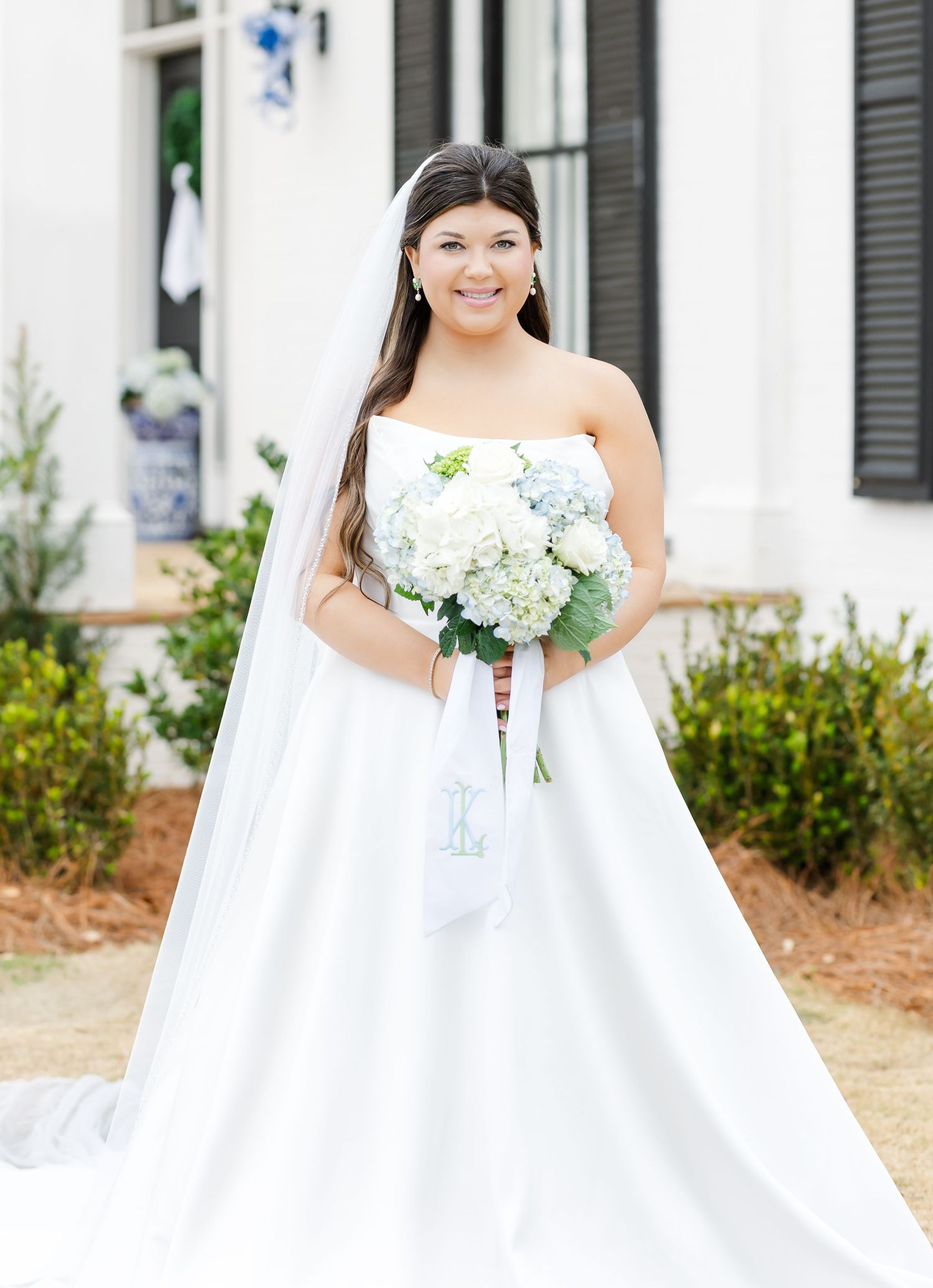 Smiling bride in strapless white ball gown holding blue and white hydrangea bouquet with veil.