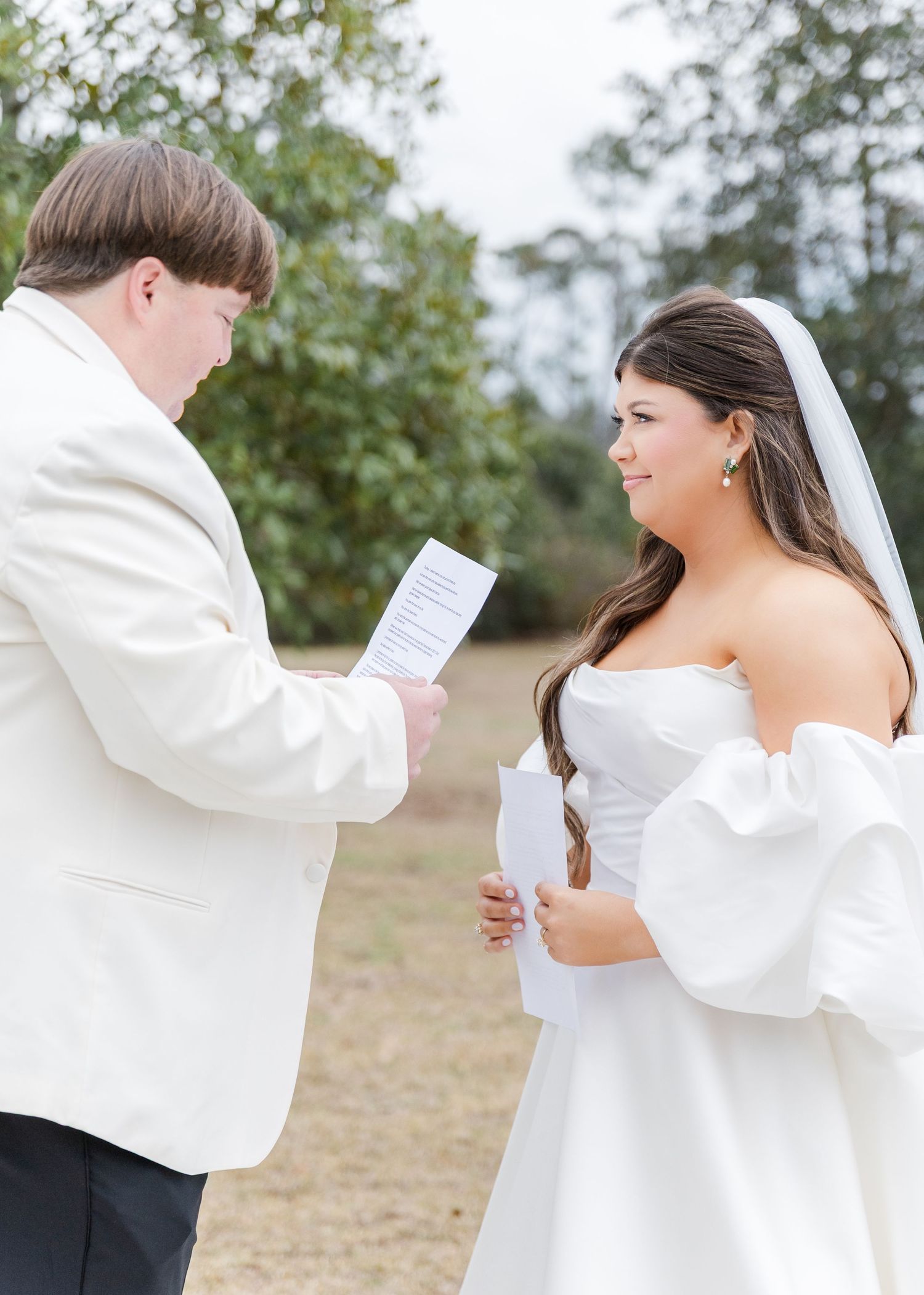 Groom in white jacket reads vows to smiling bride in strapless gown outdoors.