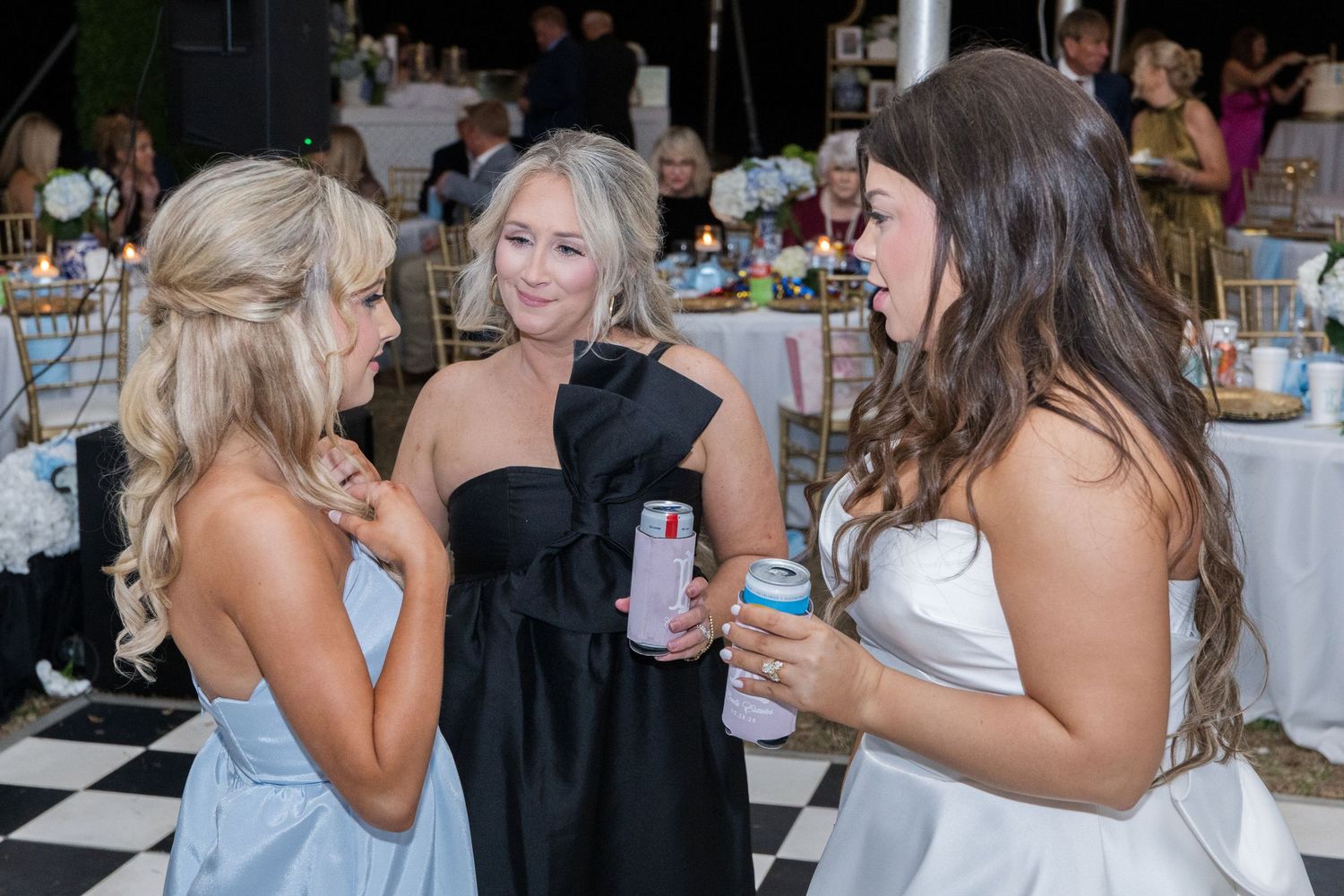 Three women in formal attire chat and hold drinks at an elegant outdoor wedding reception celebration.