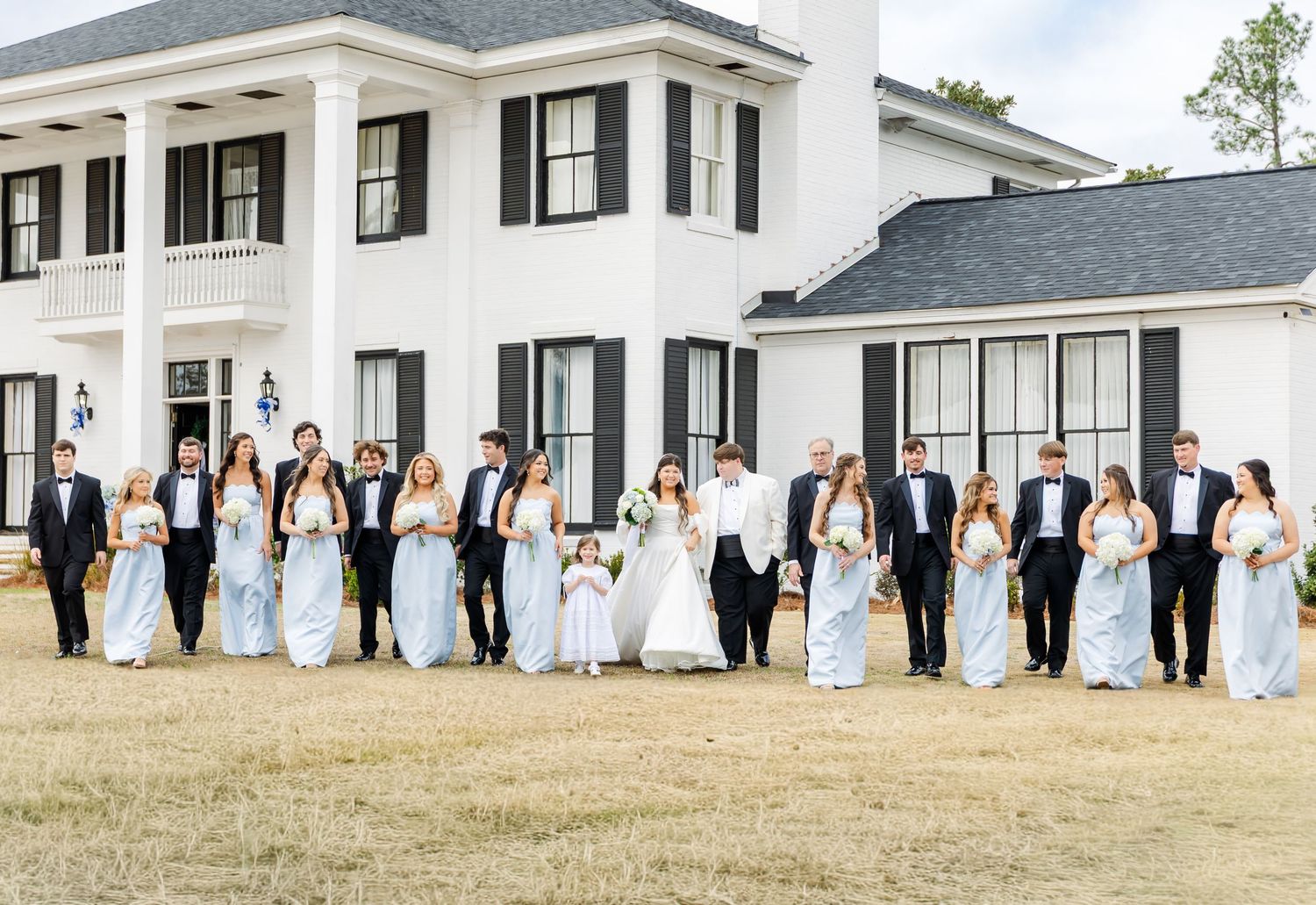 Wedding party walking across lawn in front of elegant white Southern estate with black shutters and balcony.