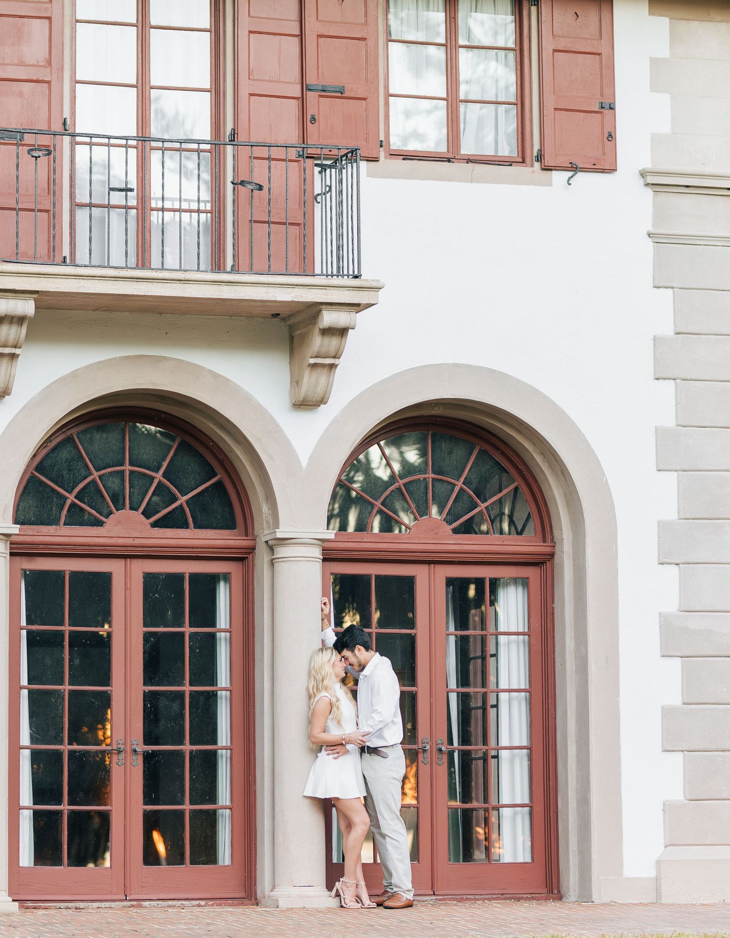 Couple shares romantic moment in front of elegant building with arched French doors during engagement photography session.