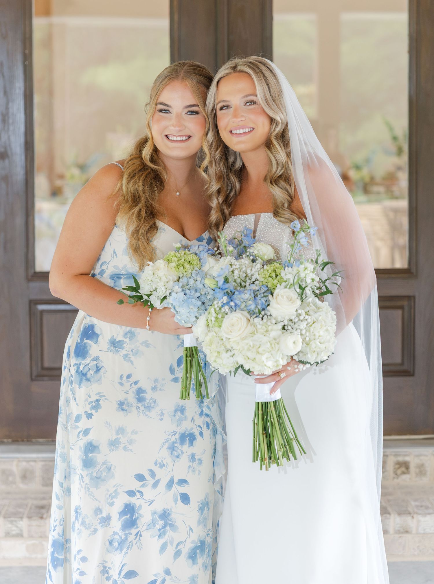 Bride in white gown with veil and bridesmaid in blue floral dress holding bouquets at wedding.