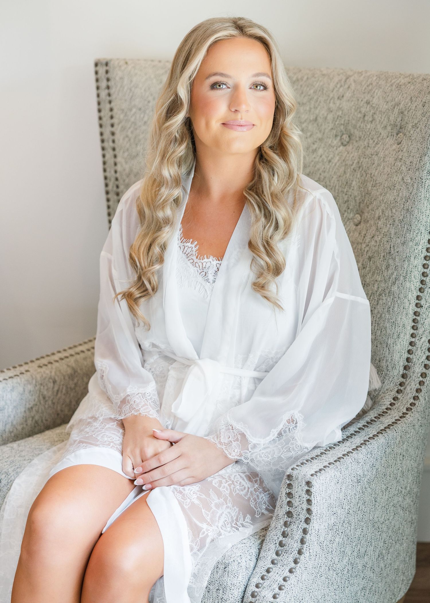 Blonde bride wearing a white lace robe sits elegantly in a tufted gray chair during wedding morning preparations.