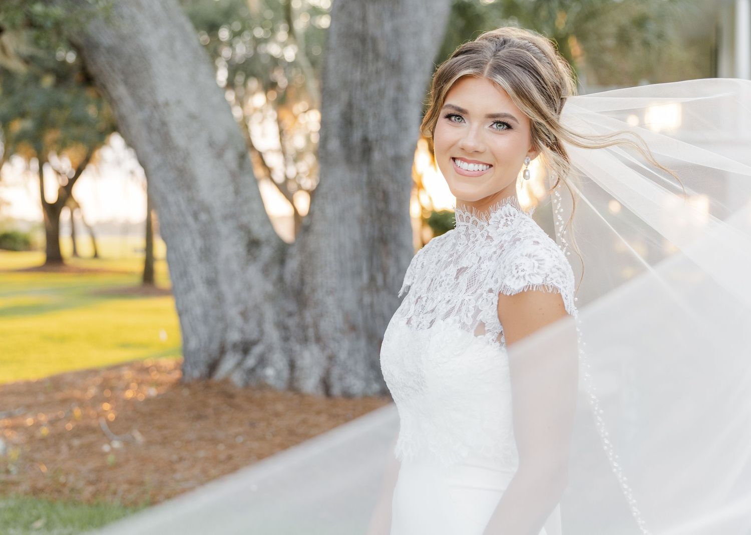 Bride in lace high-neck wedding gown with flowing veil smiling outdoors near large oak tree.
