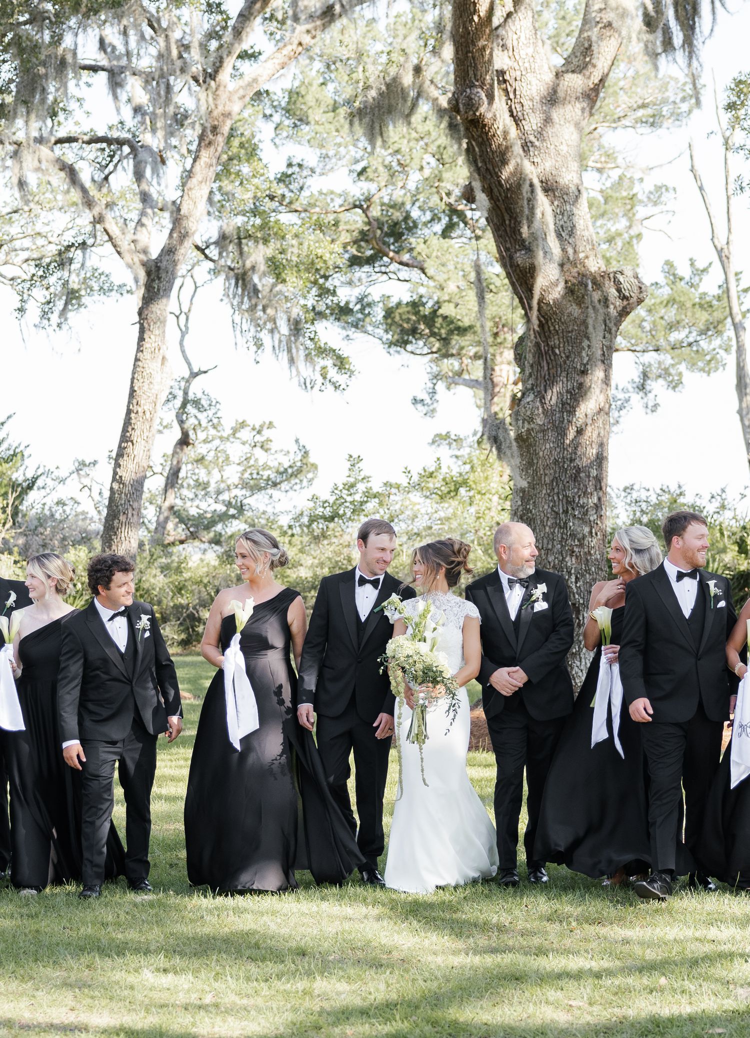 Wedding party in black formal attire walks outdoors under moss-draped trees, bride in white gown holding bouquet.