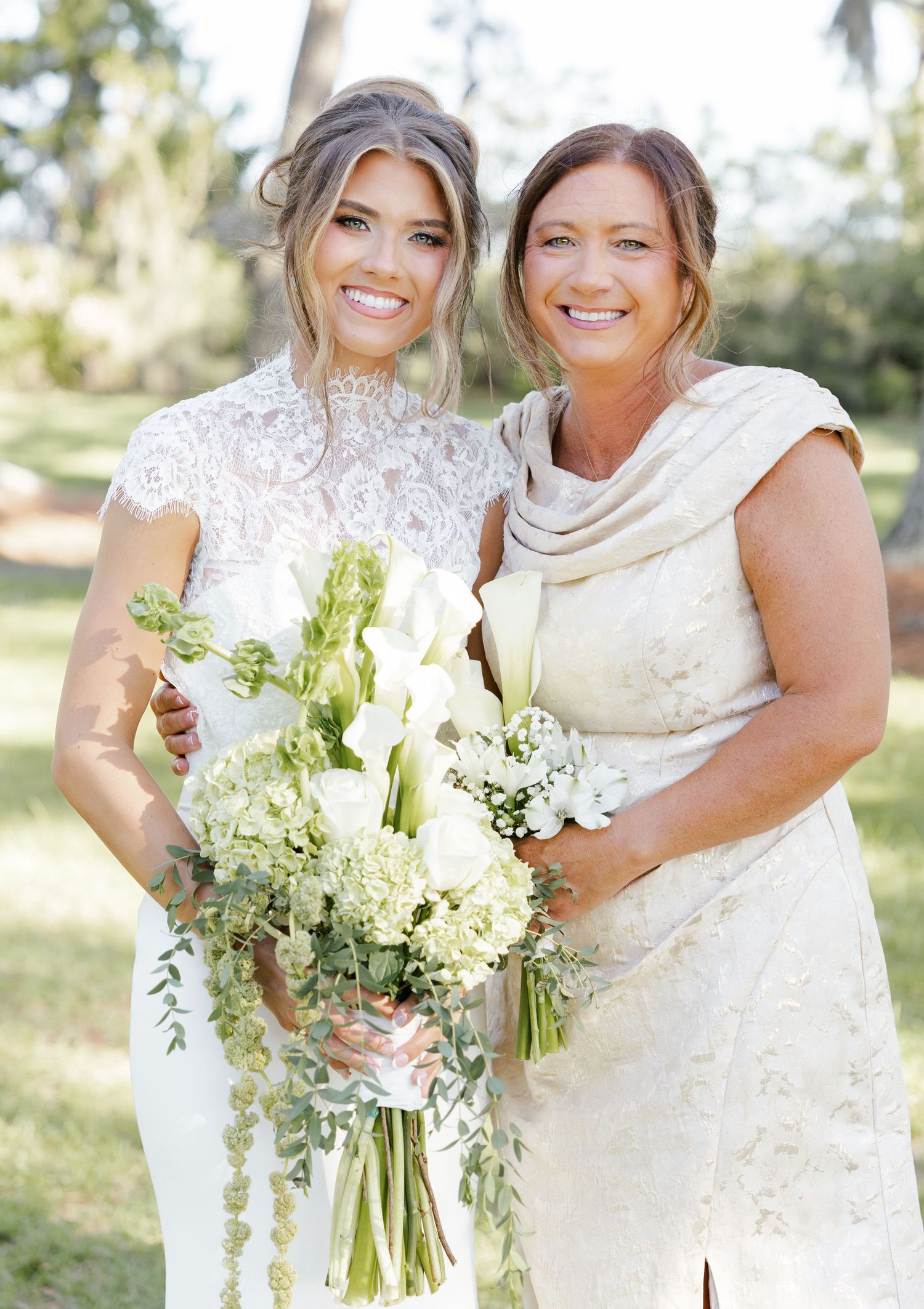 Bride in white lace gown holds green and white bouquet beside woman in beige dress outdoors.