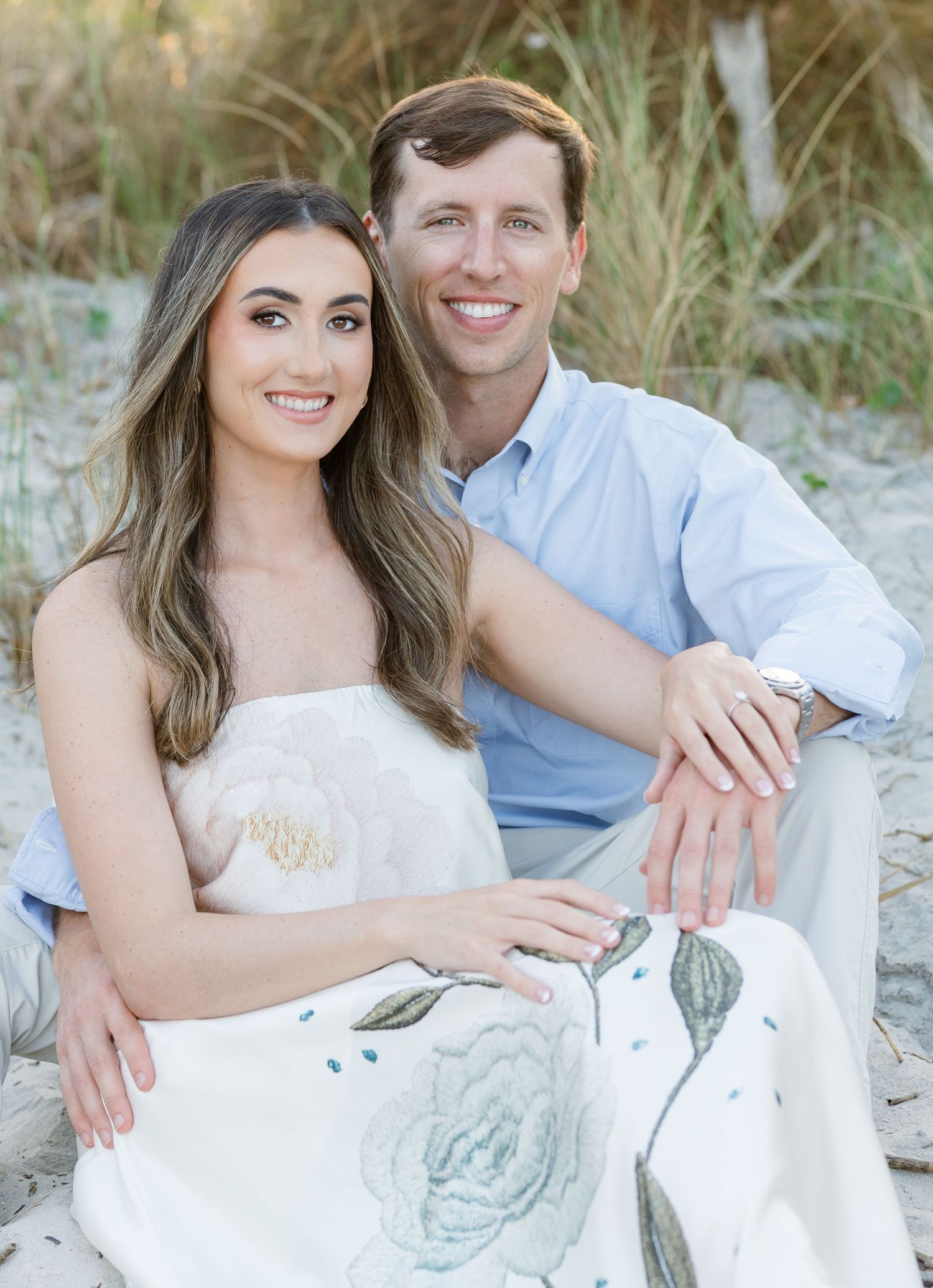 A smiling couple poses together on a sandy beach, the woman in a floral dress and man in a light blue shirt.