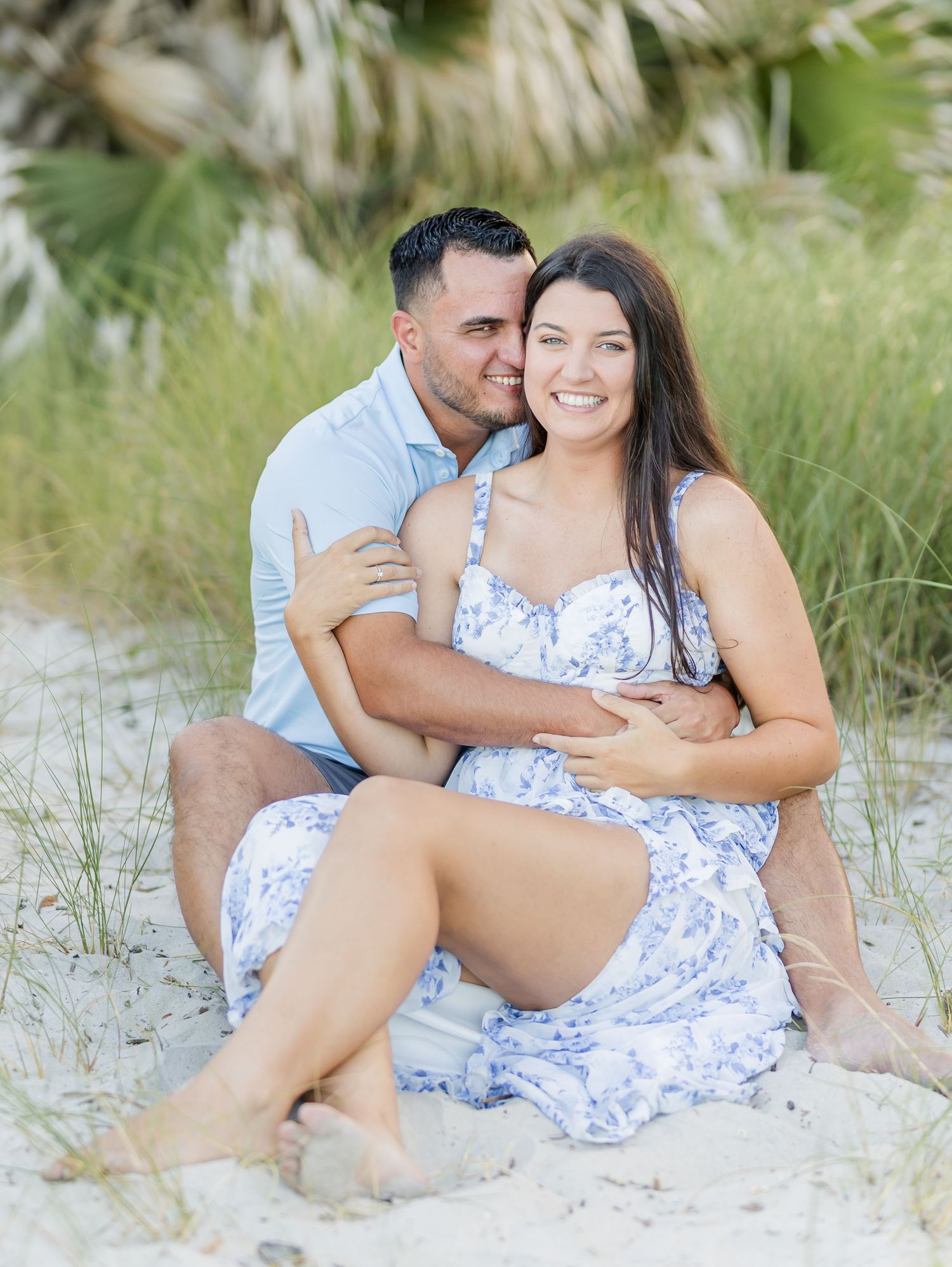 A couple in matching blue and white outfits share intimate moments on a sandy beach with sea grass in the background.