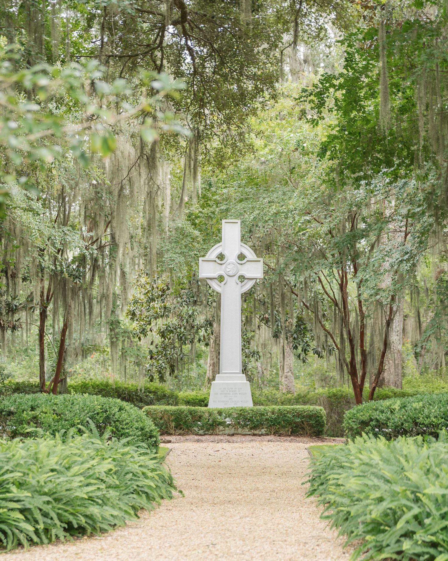Stone cross memorial markers stand along a winding gravel path surrounded by lush ferns and Spanish moss draped trees.