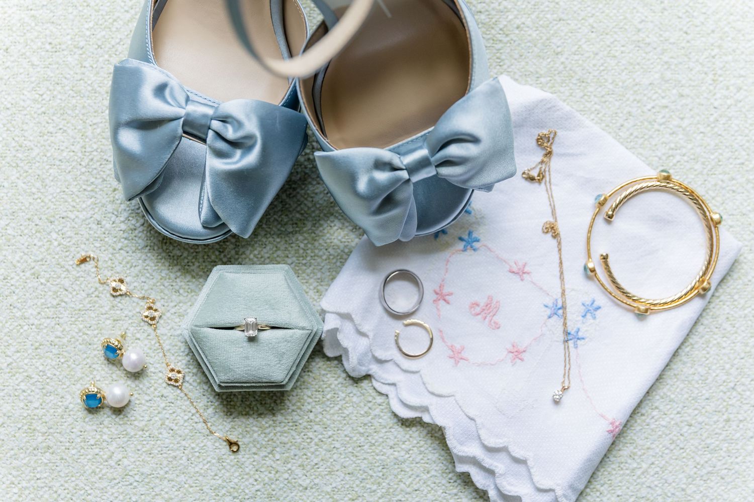Wedding accessories including blue bow heels, rings, and jewelry on white fabric.