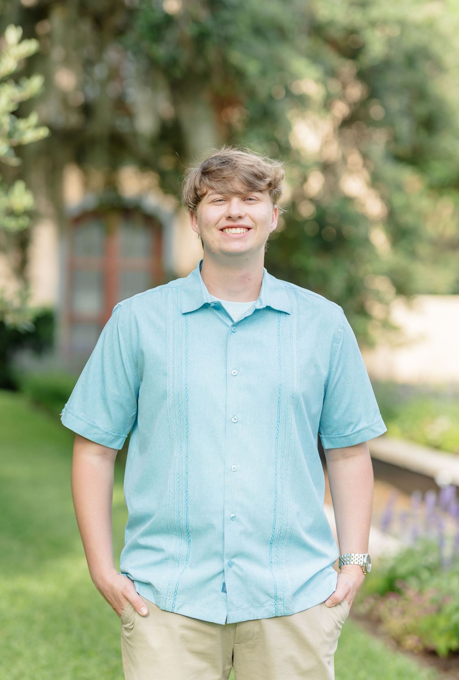 A person wearing a light blue short-sleeve button-up shirt and khaki pants poses in front of a leafy garden setting.