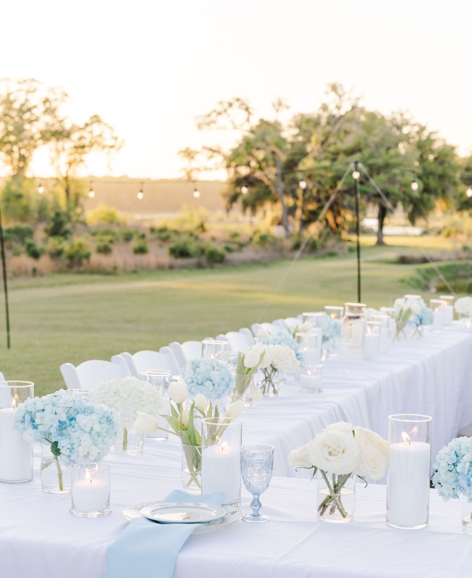 Elegant outdoor wedding reception table with white linens, blue hydrangeas, candles, and string lights at golden hour.