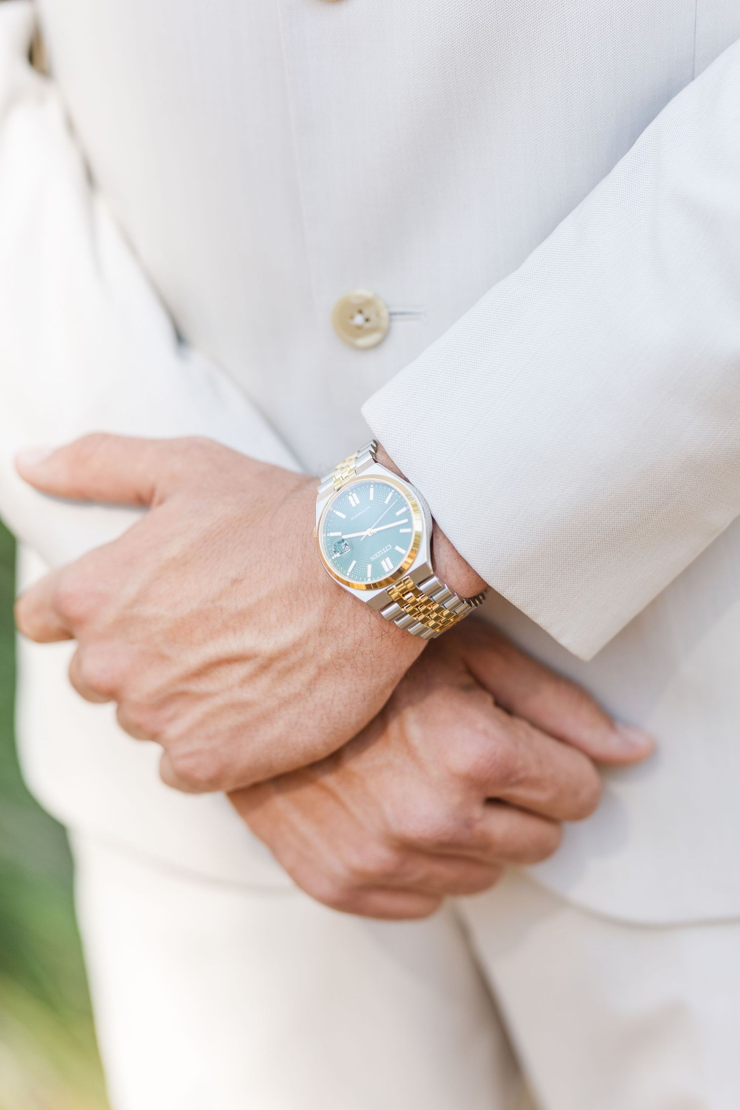 Close-up detail of hands wearing a white suit jacket sleeve with a blue-faced watch on the wrist.
