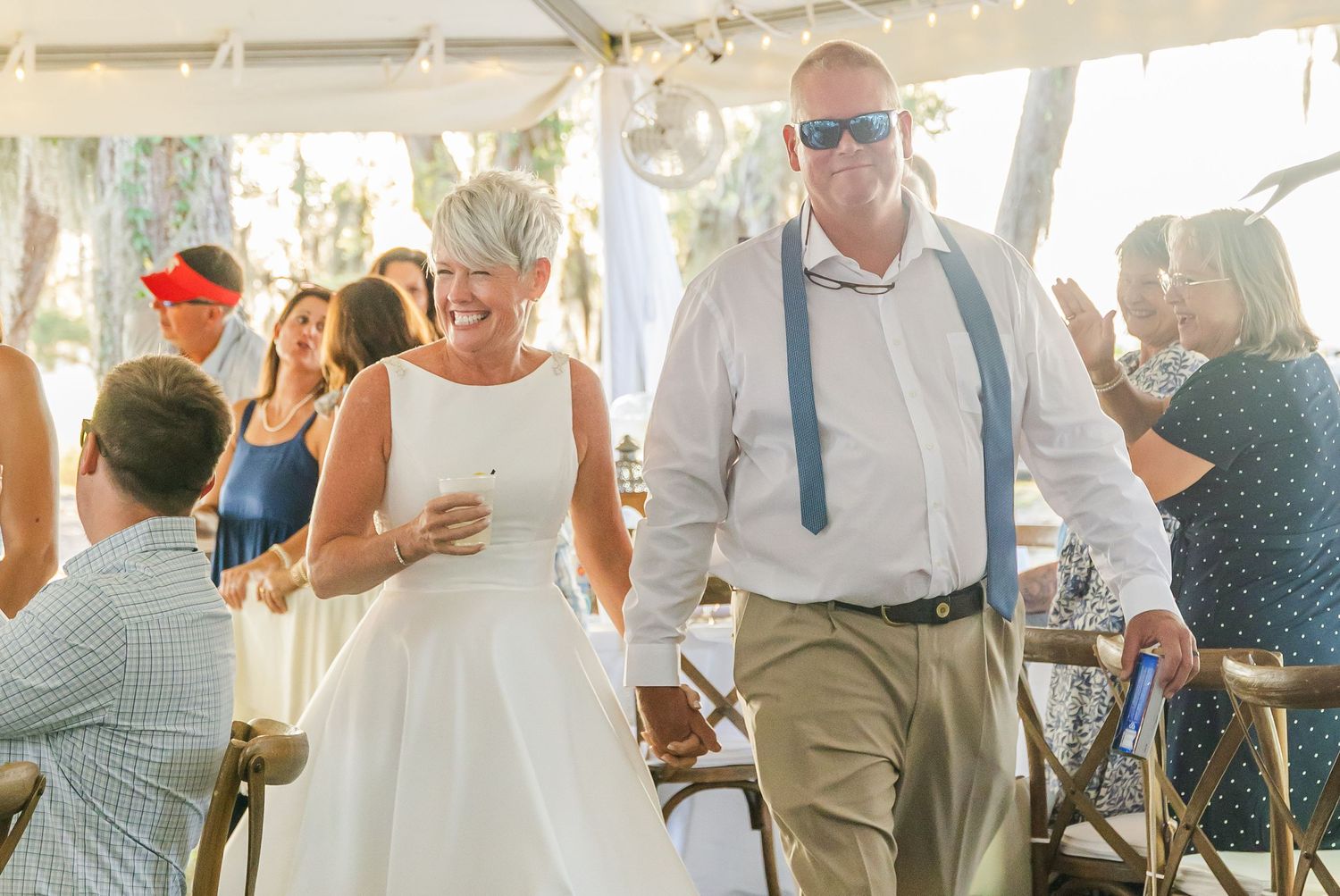 Wedding guests mingle and socialize during an outdoor reception under a white tent on a sunny day.