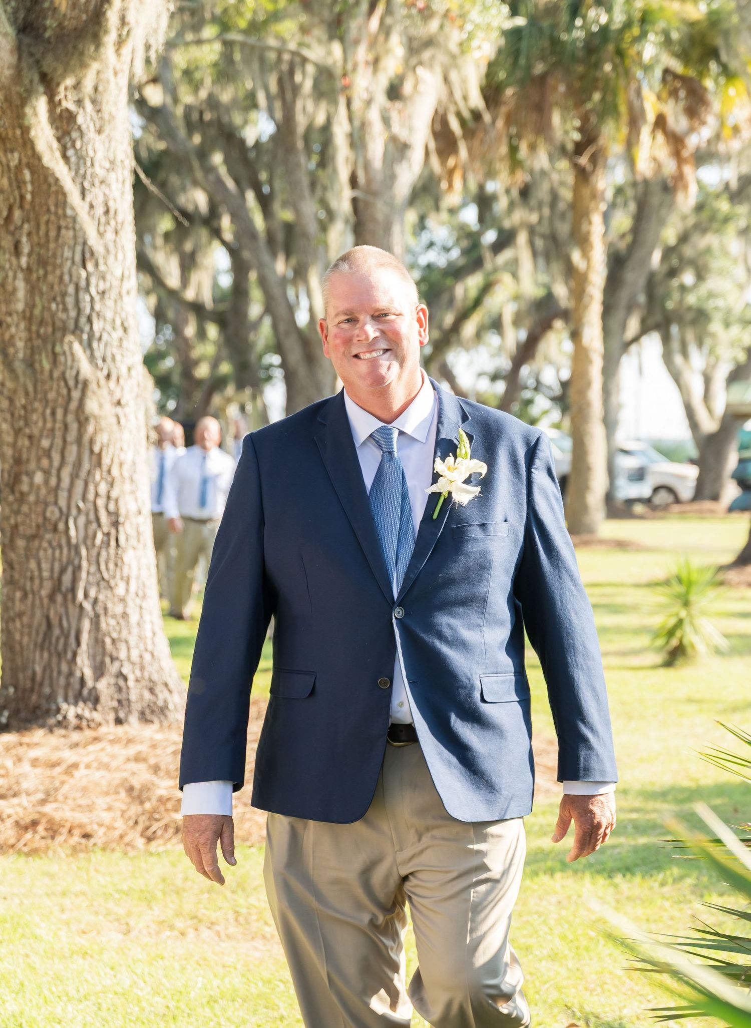 Someone wearing a navy suit jacket with khaki pants stands smiling outdoors among Spanish moss-draped trees.