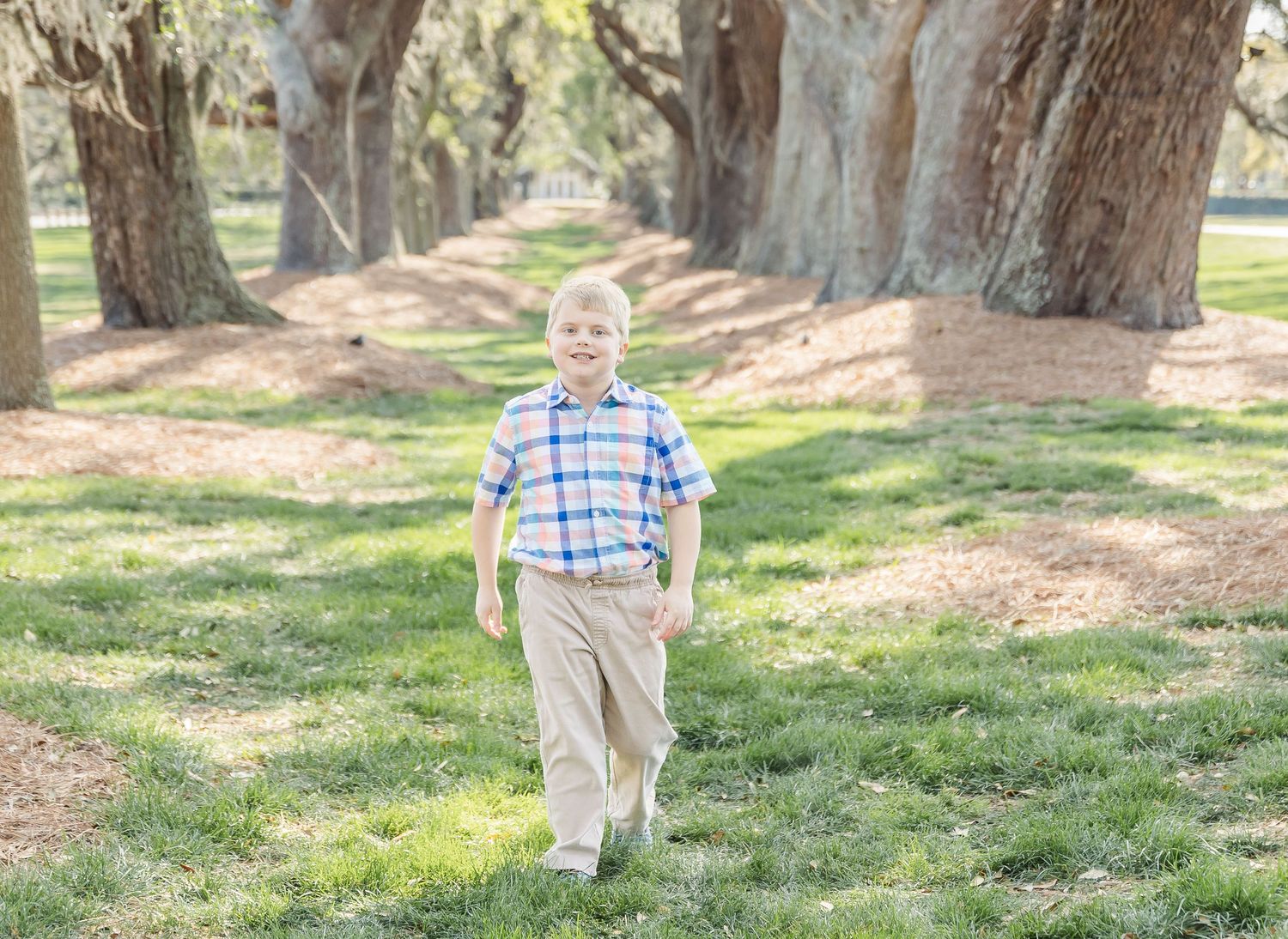 Young boy in plaid shirt walks confidently down oak-lined path during outdoor photo session.