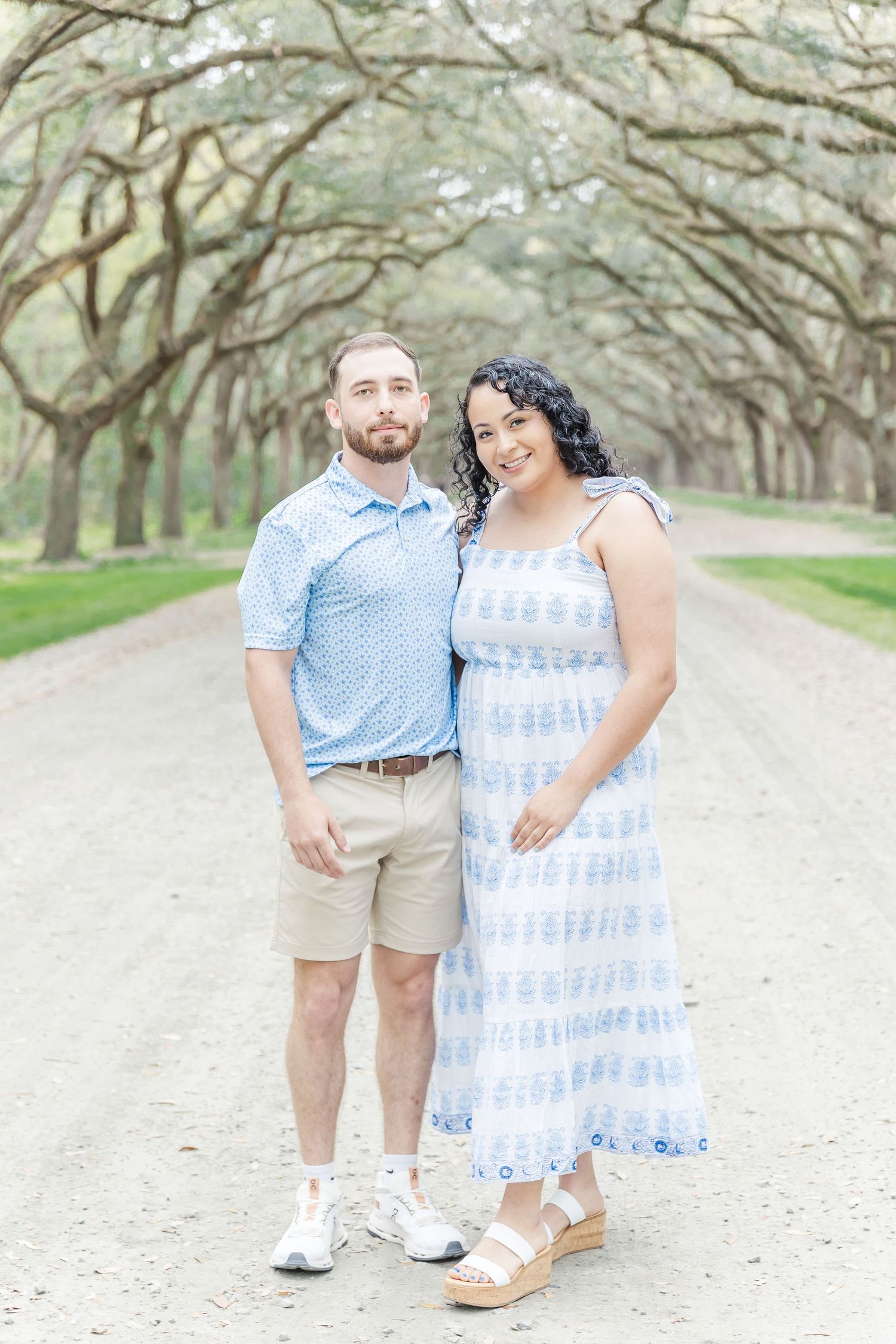 Couple in coordinating blue and white outfits pose on scenic oak-lined pathway.