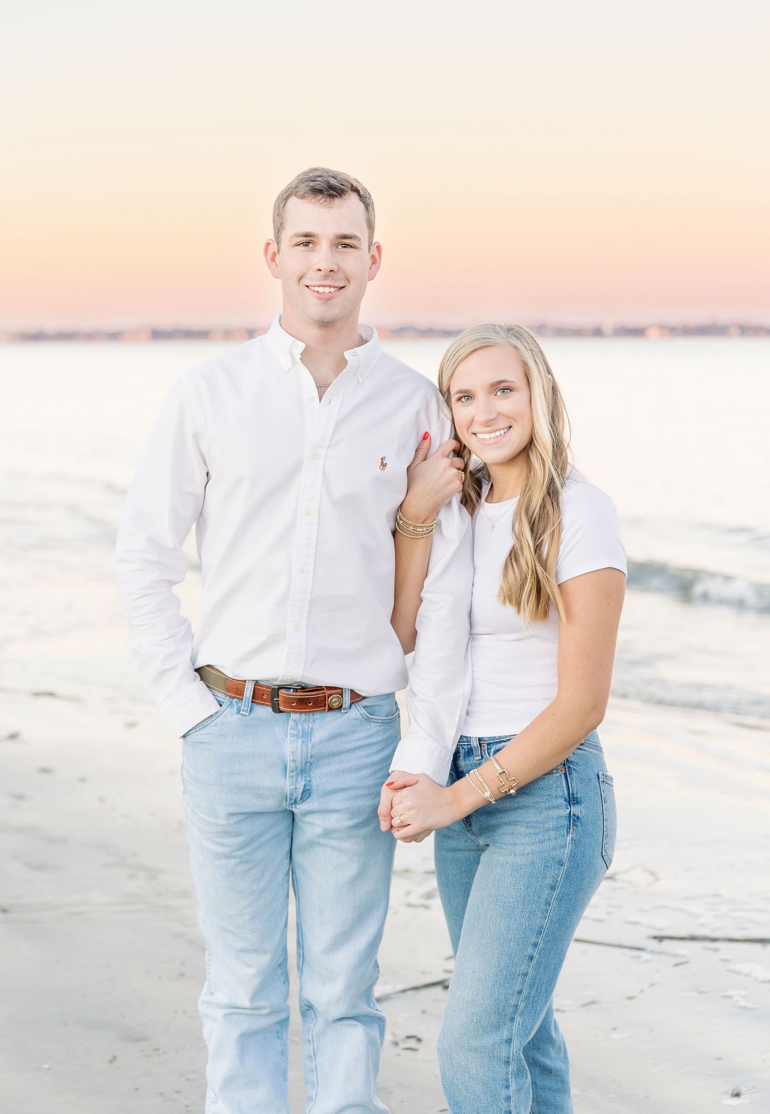 Couple in coordinated white tops and jeans smiling on a beach during golden hour.