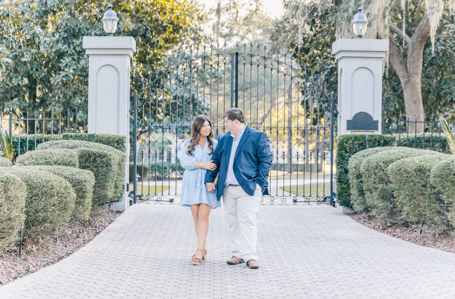 Couple standing in front of grand white pillars and iron gates on a brick pathway.