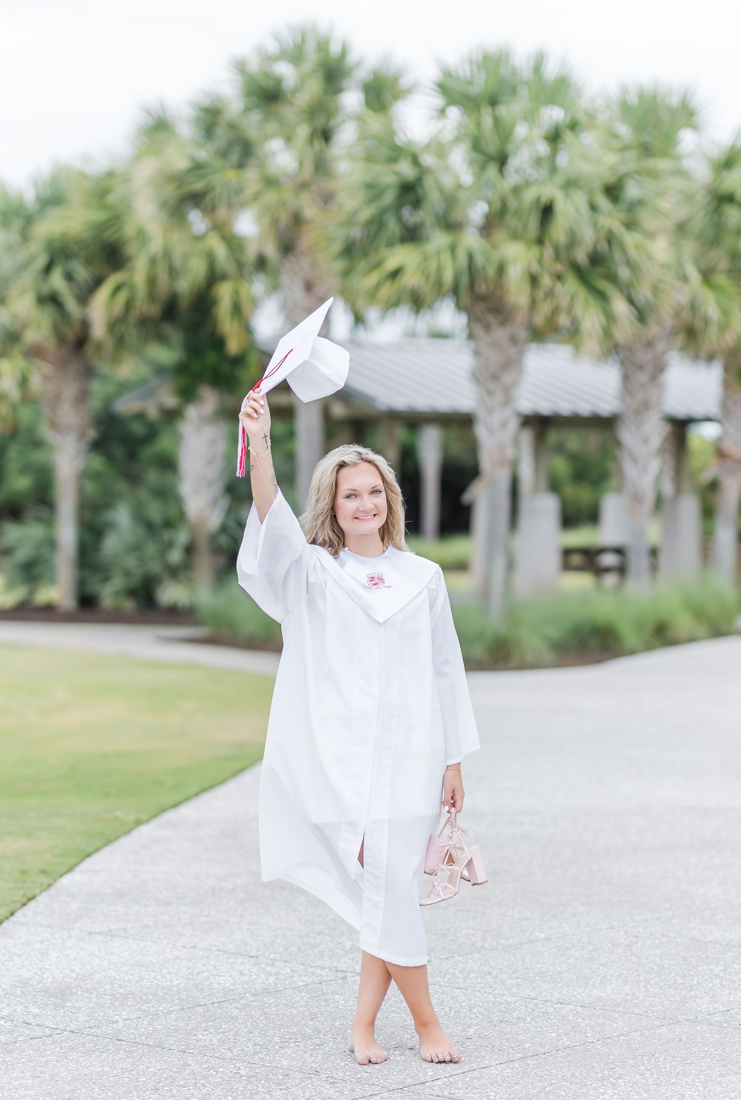 Graduate in white gown holds cap up while standing barefoot on path near palm trees.