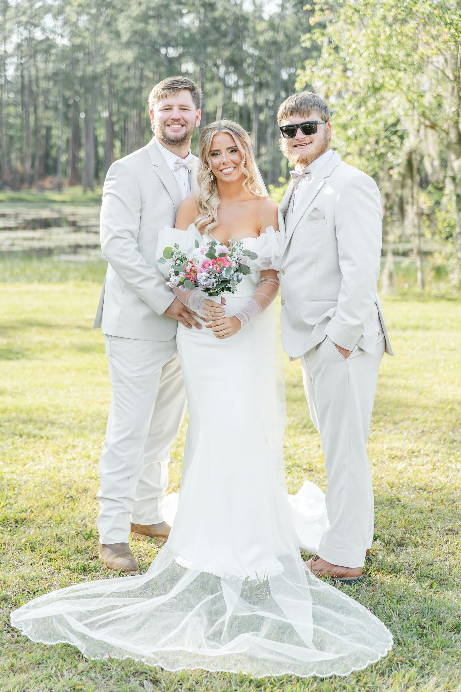 Three people in white formal wear share moment with floral bouquet outdoors.