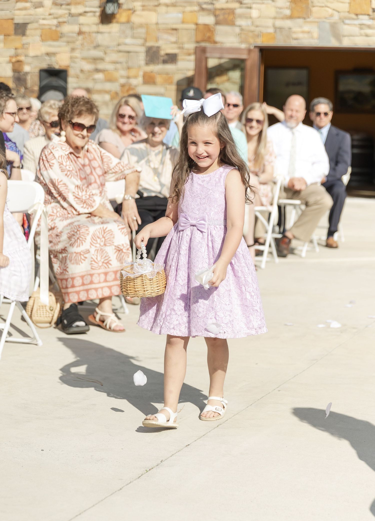 Flower girl in lavender dress carrying basket at outdoor ceremony with stone building backdrop.