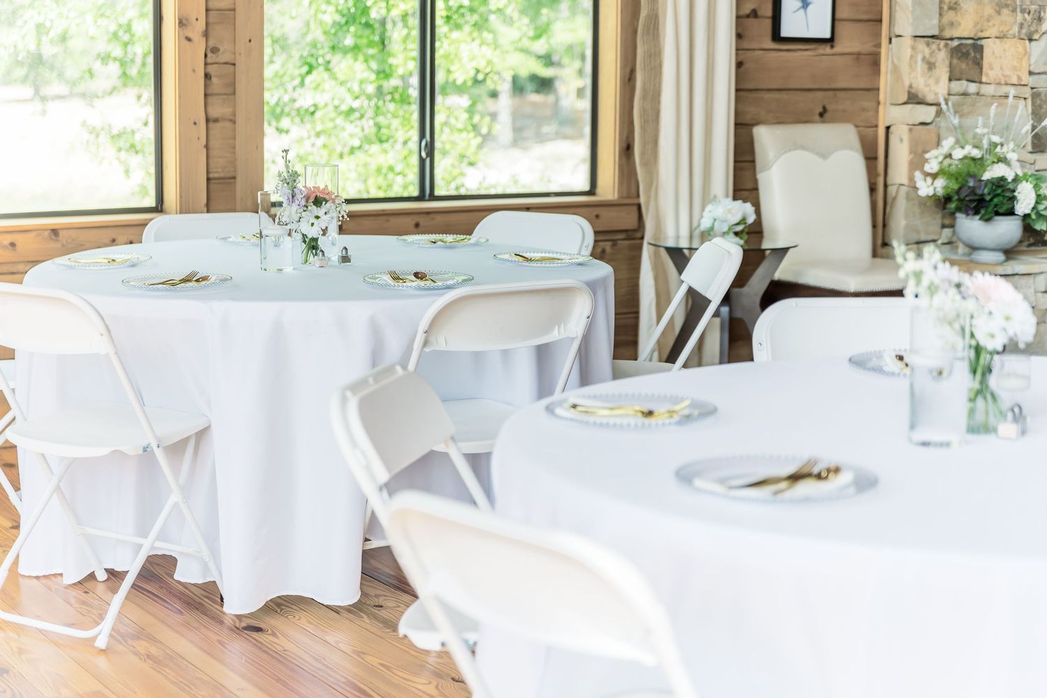 Elegant white table settings with folding chairs in a sunlit rustic venue with wooden walls.