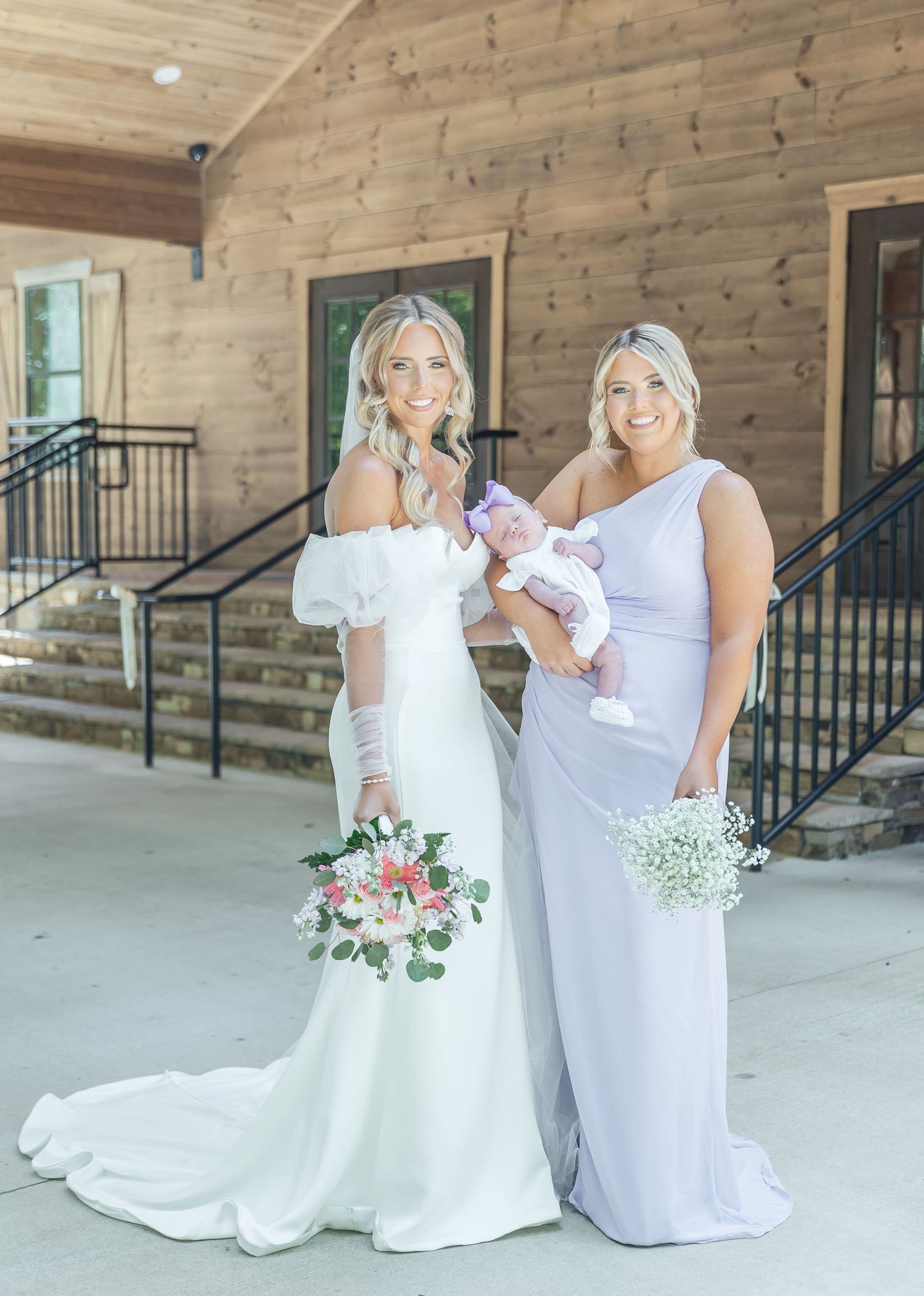 Wedding party members pose with an infant on rustic porch with floral arrangements.