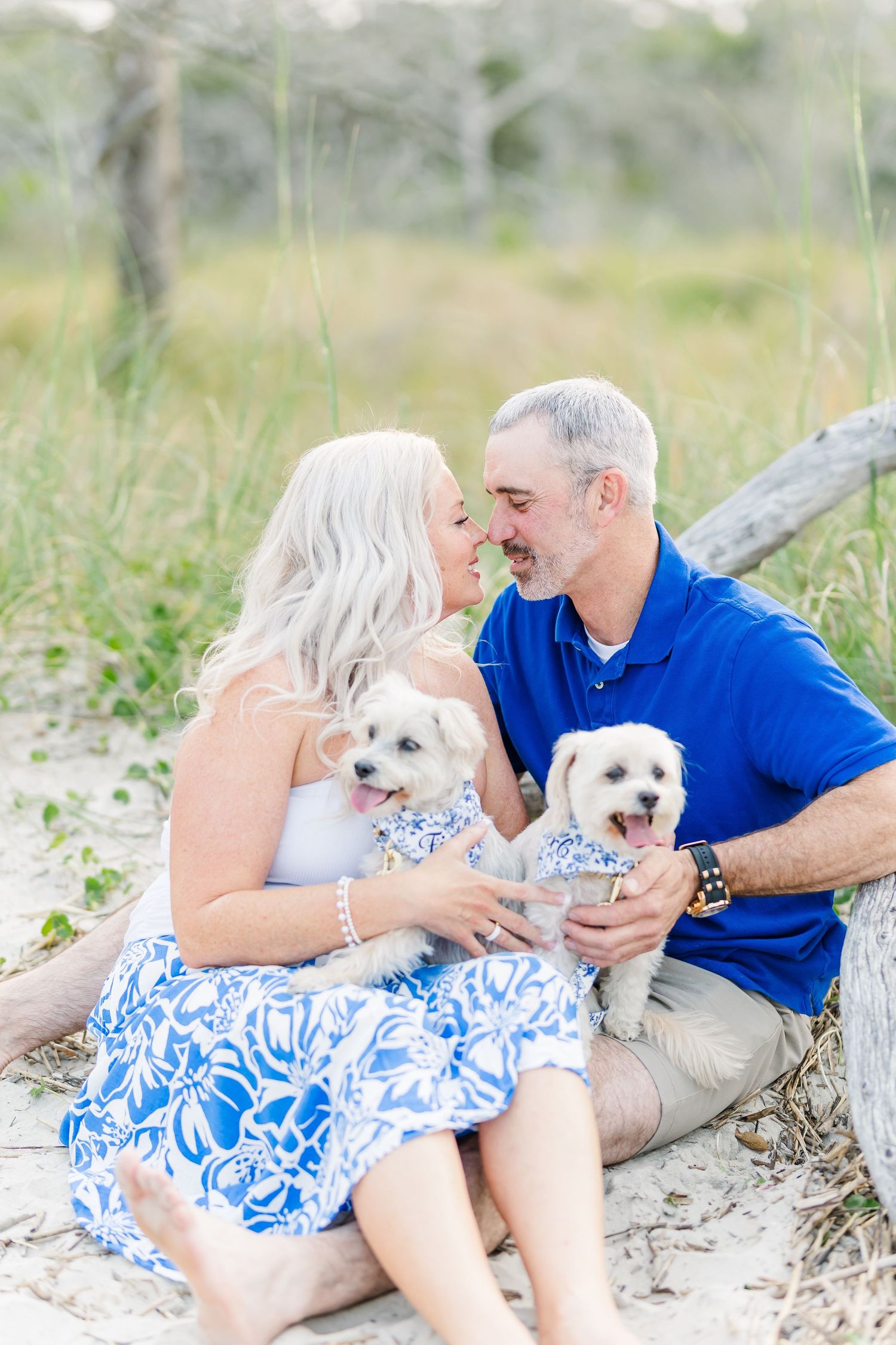 A couple sits with their two small white dogs on a beach with driftwood.