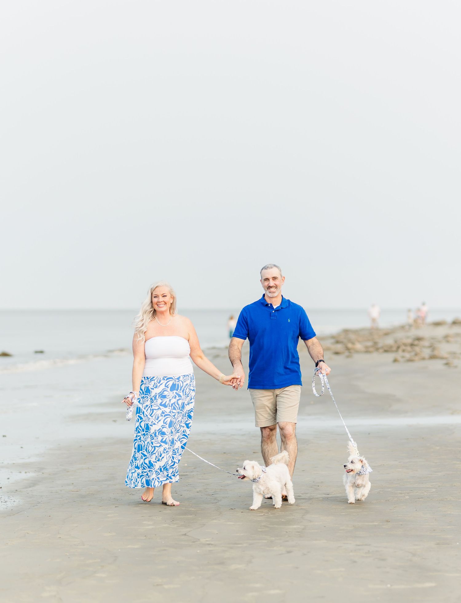 A couple walks their two small white dogs along the shoreline at the beach on a sunny day.
