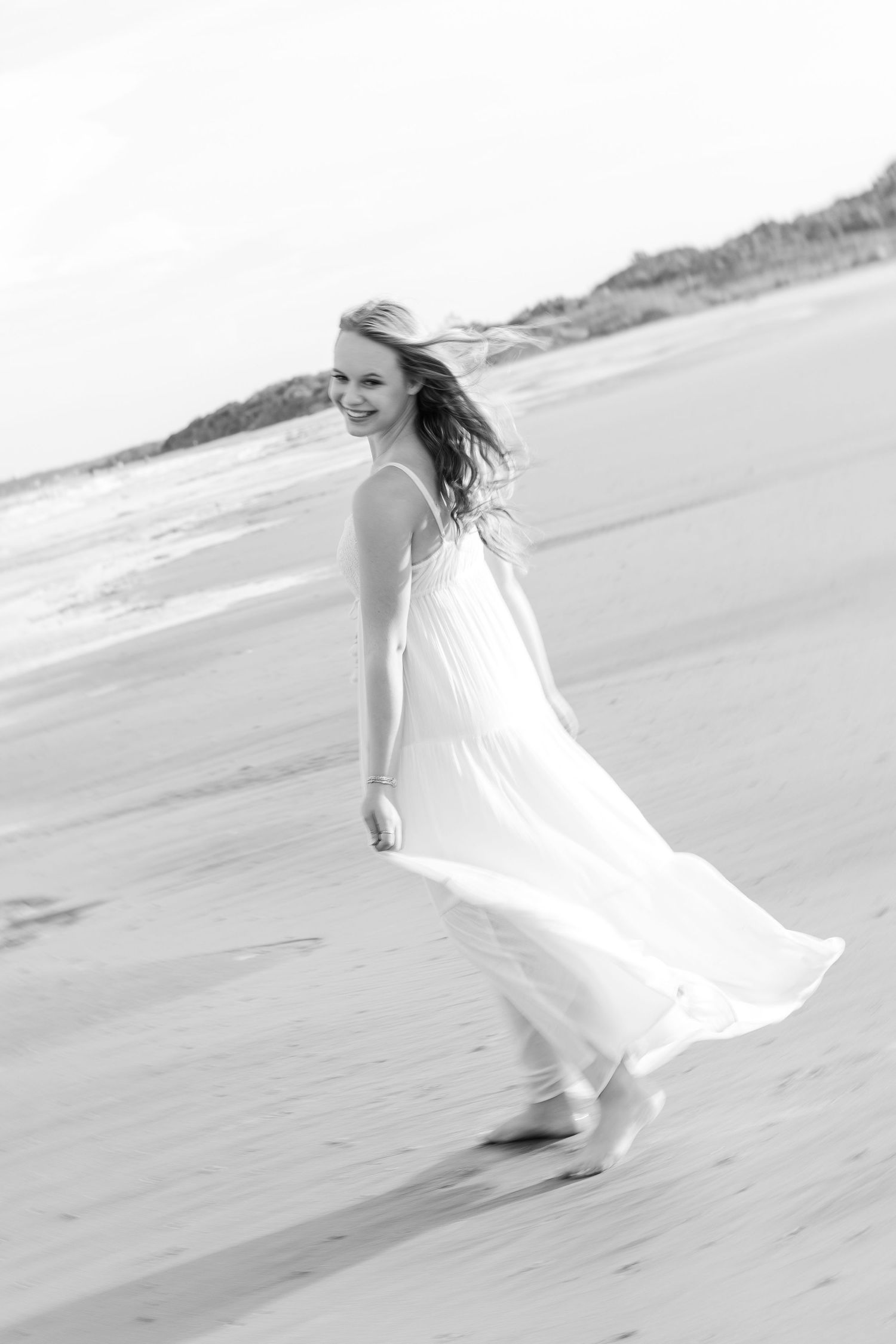 A flowing white dress catches the ocean breeze as someone twirls on a sandy beach in black and white.