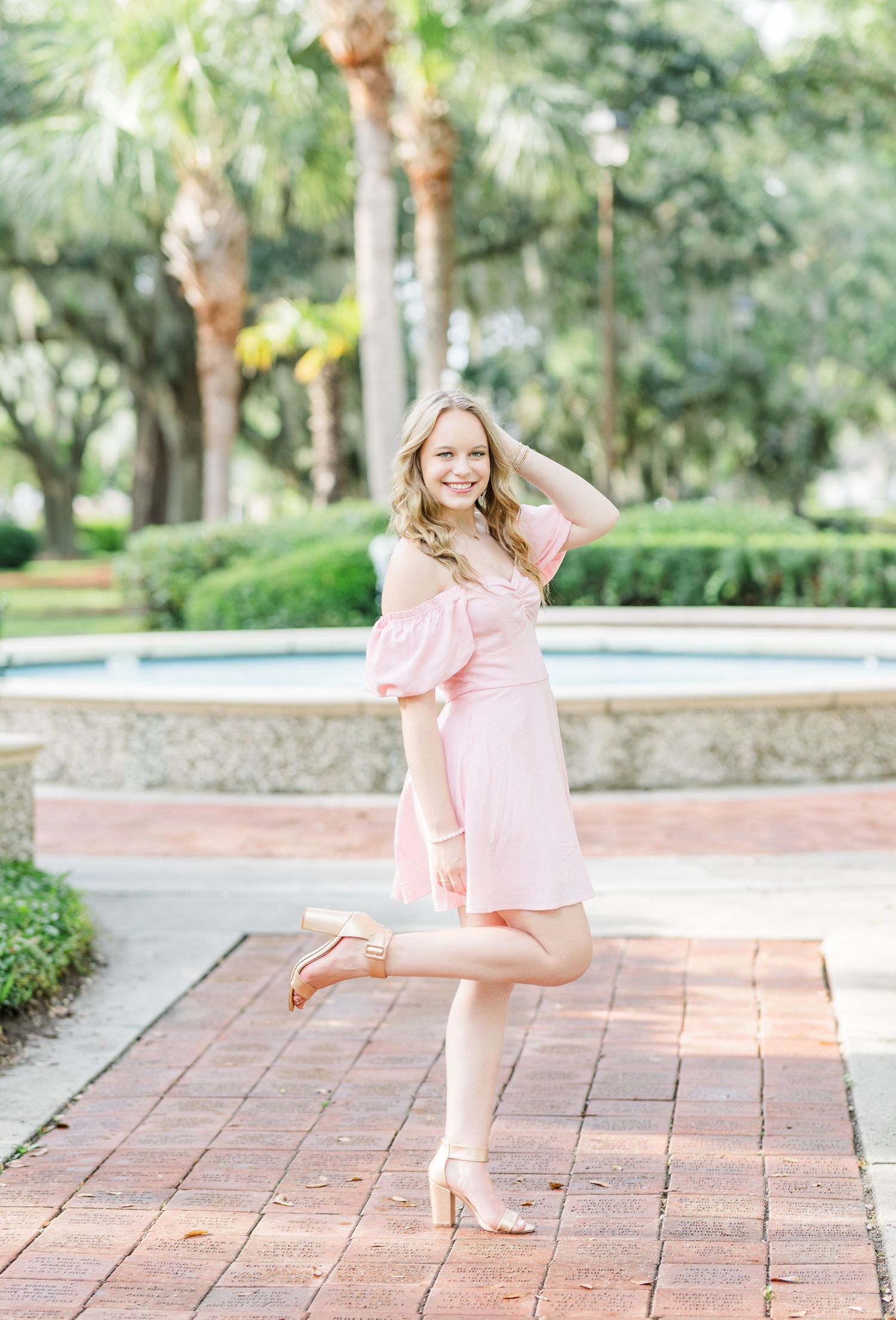 Someone wearing a pink dress strikes a playful pose on brick pavers in a garden setting.