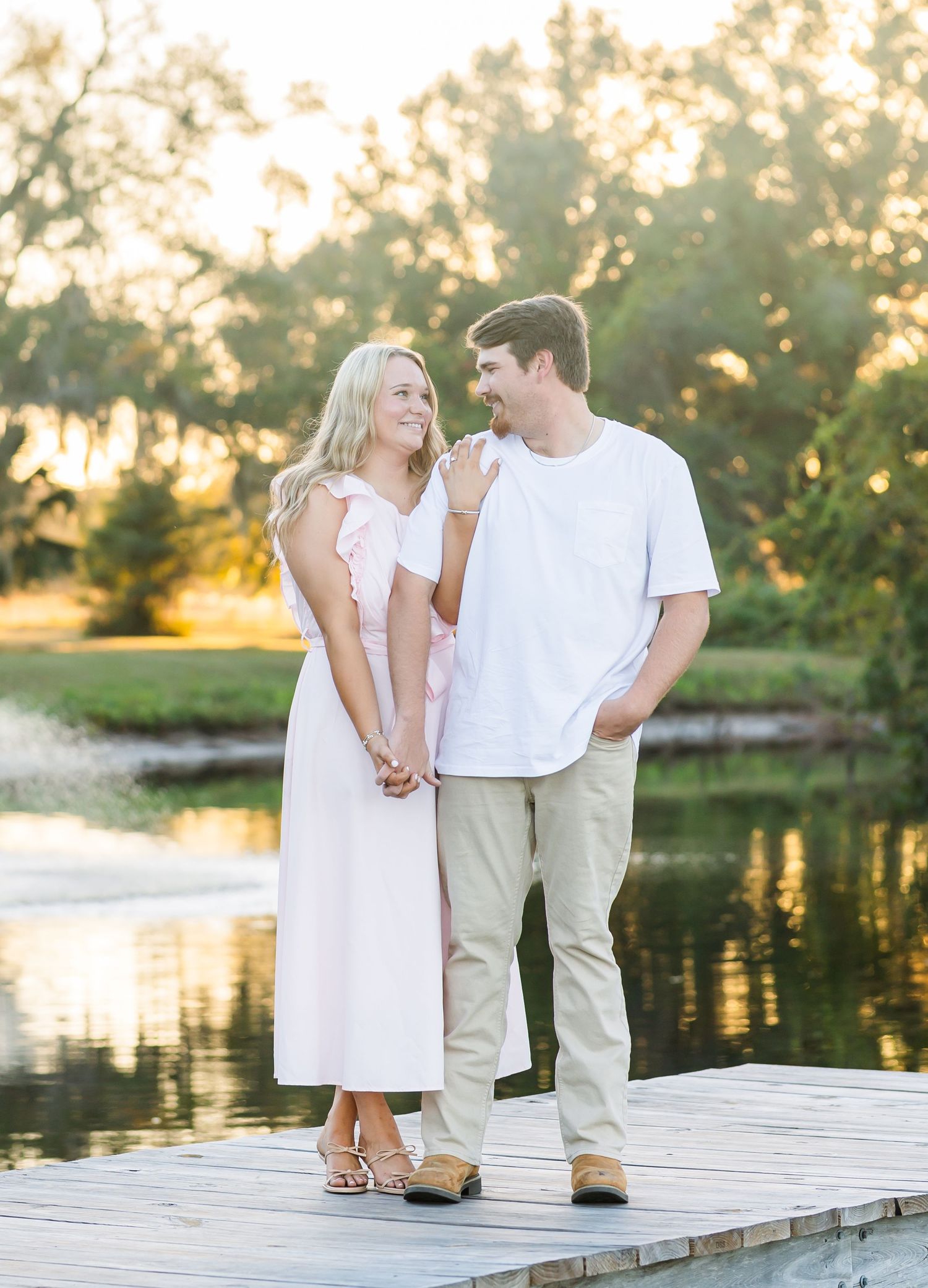 Romantic couple stands together on wooden dock by serene lake at golden sunset hour.