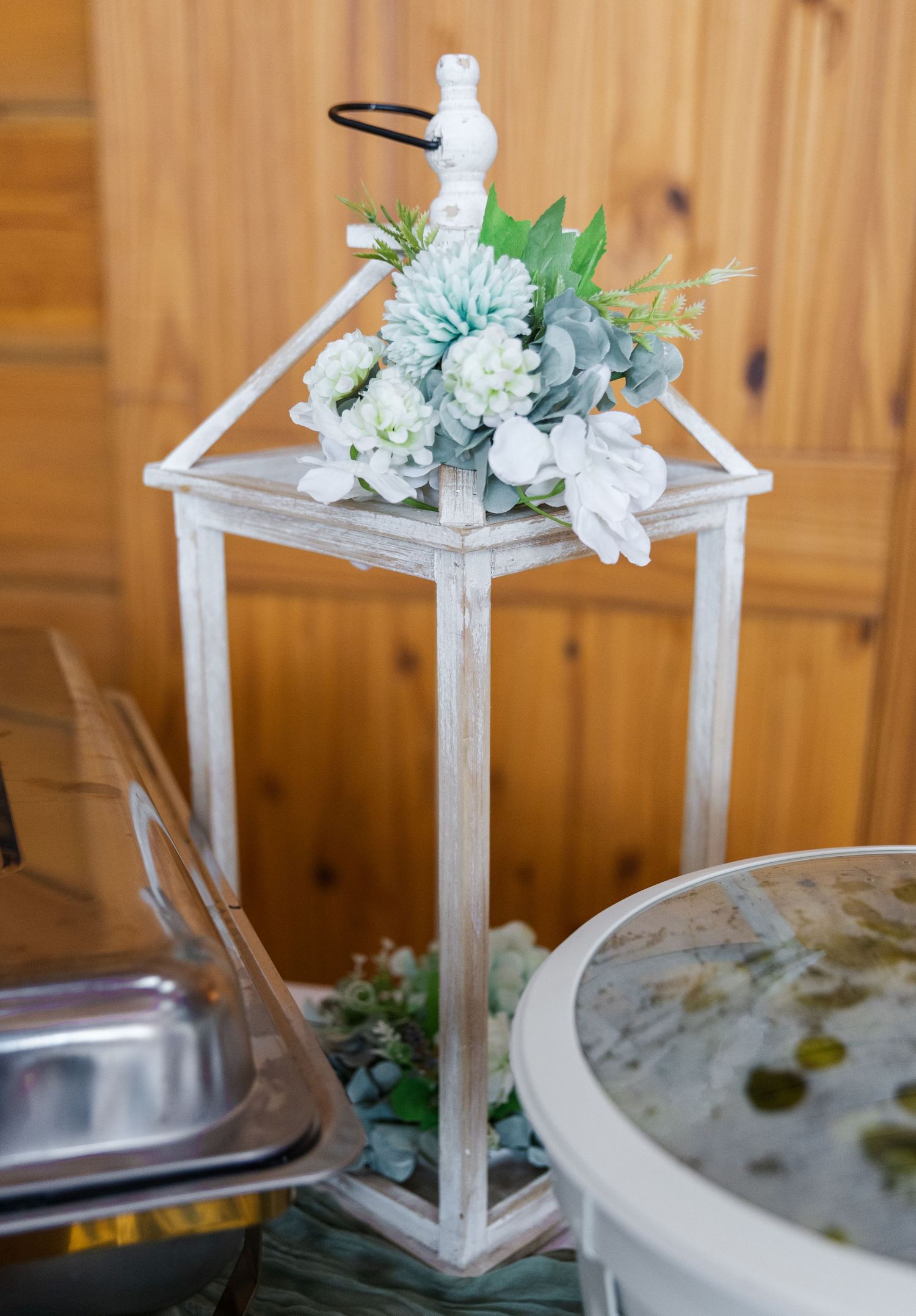 A white wooden lantern decorated with light blue and white floral arrangements sits beside catering dishes.