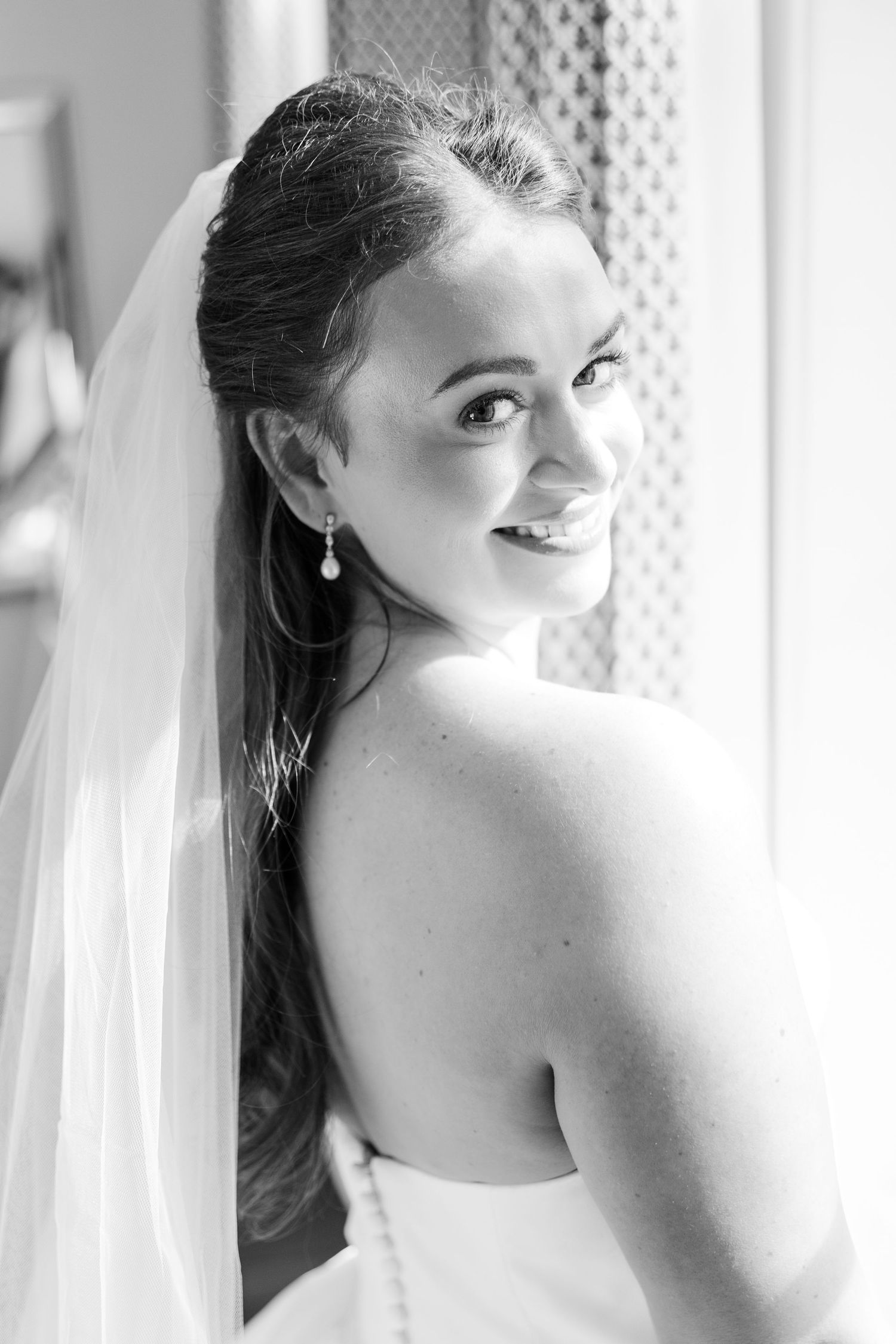 Black and white bridal portrait showing a bride in a veil looking over her shoulder with a joyful expression.