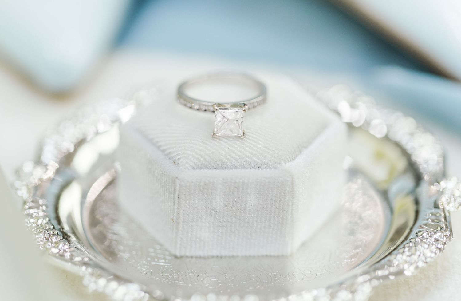 A sparkling diamond engagement ring sits atop a white velvet cushion on an ornate silver tray.
