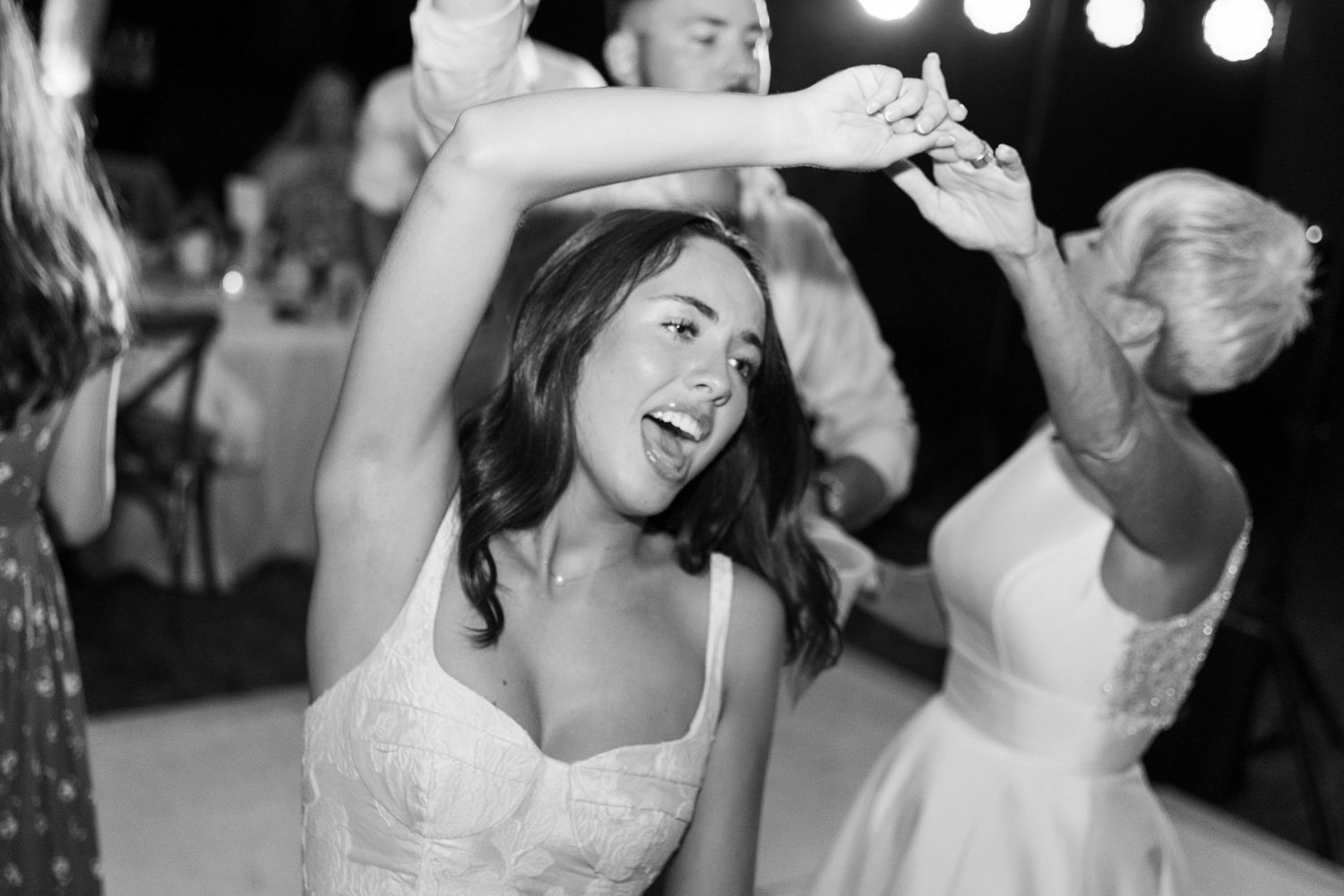 Black and white shot of joyful dancing couples at wedding celebration under string lights.