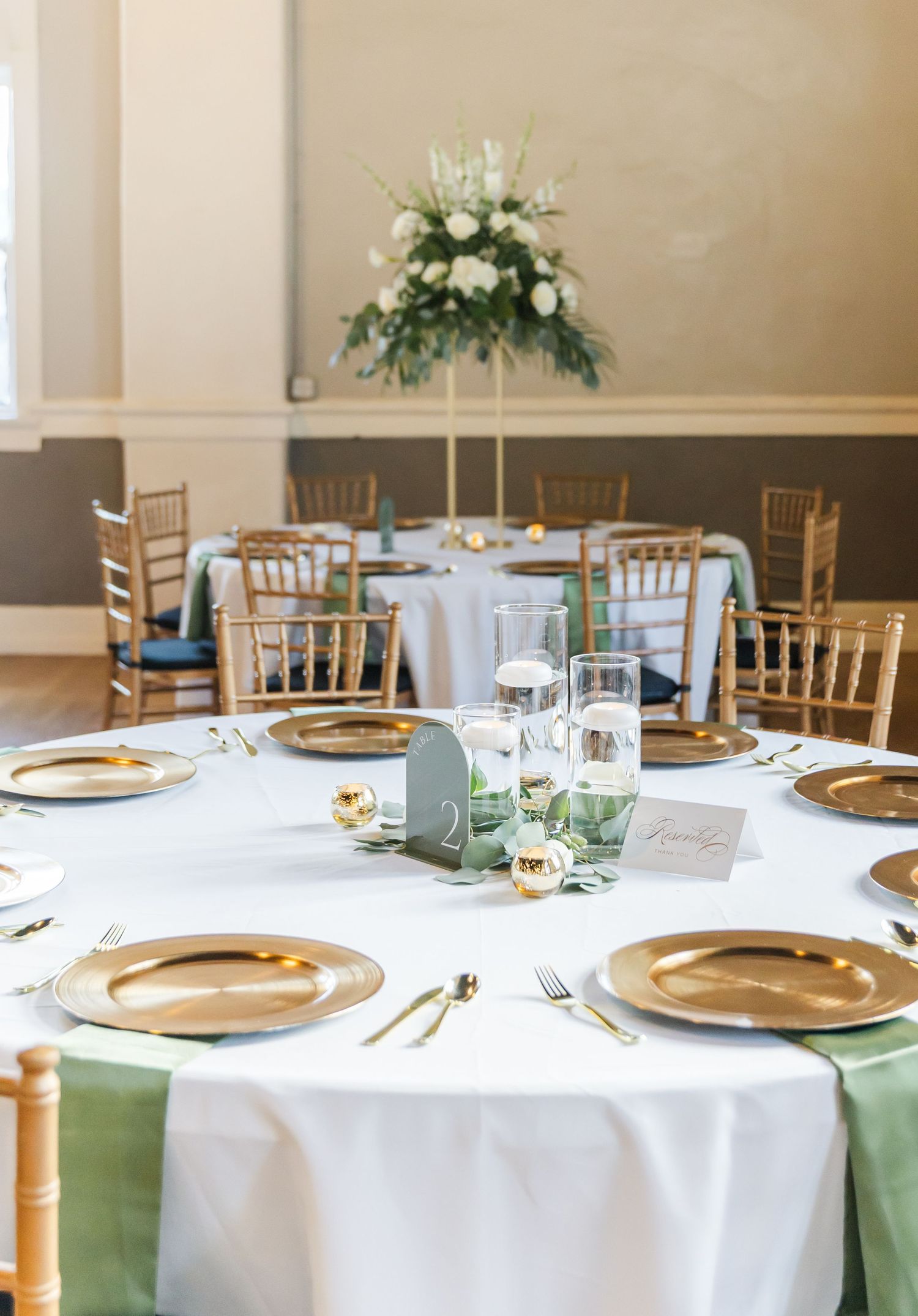 Elegant table settings featuring gold charger plates, sage green napkins, and white floral centerpieces.