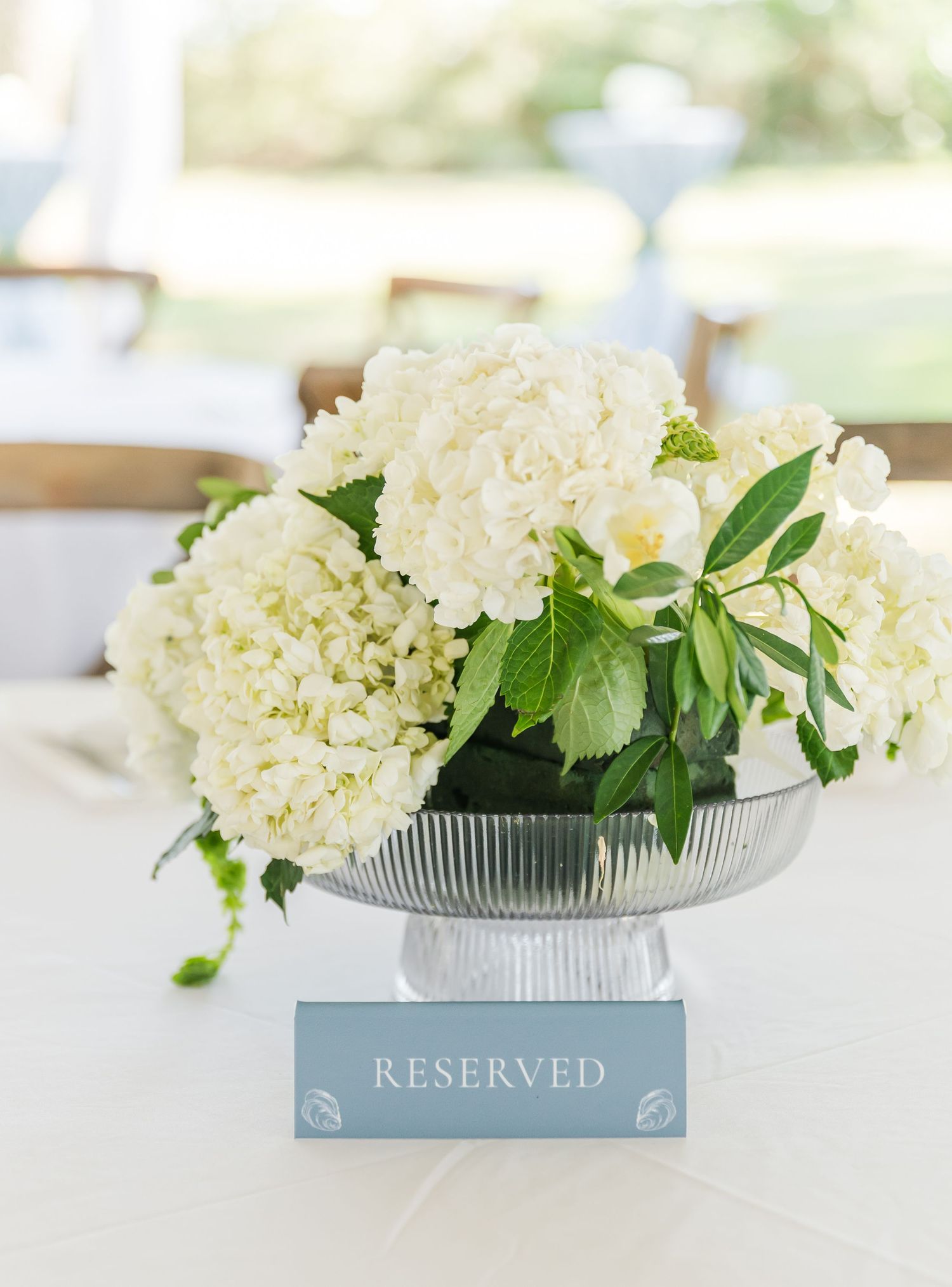 White hydrangea floral arrangement in a silver bowl with a reserved sign on a table setting.