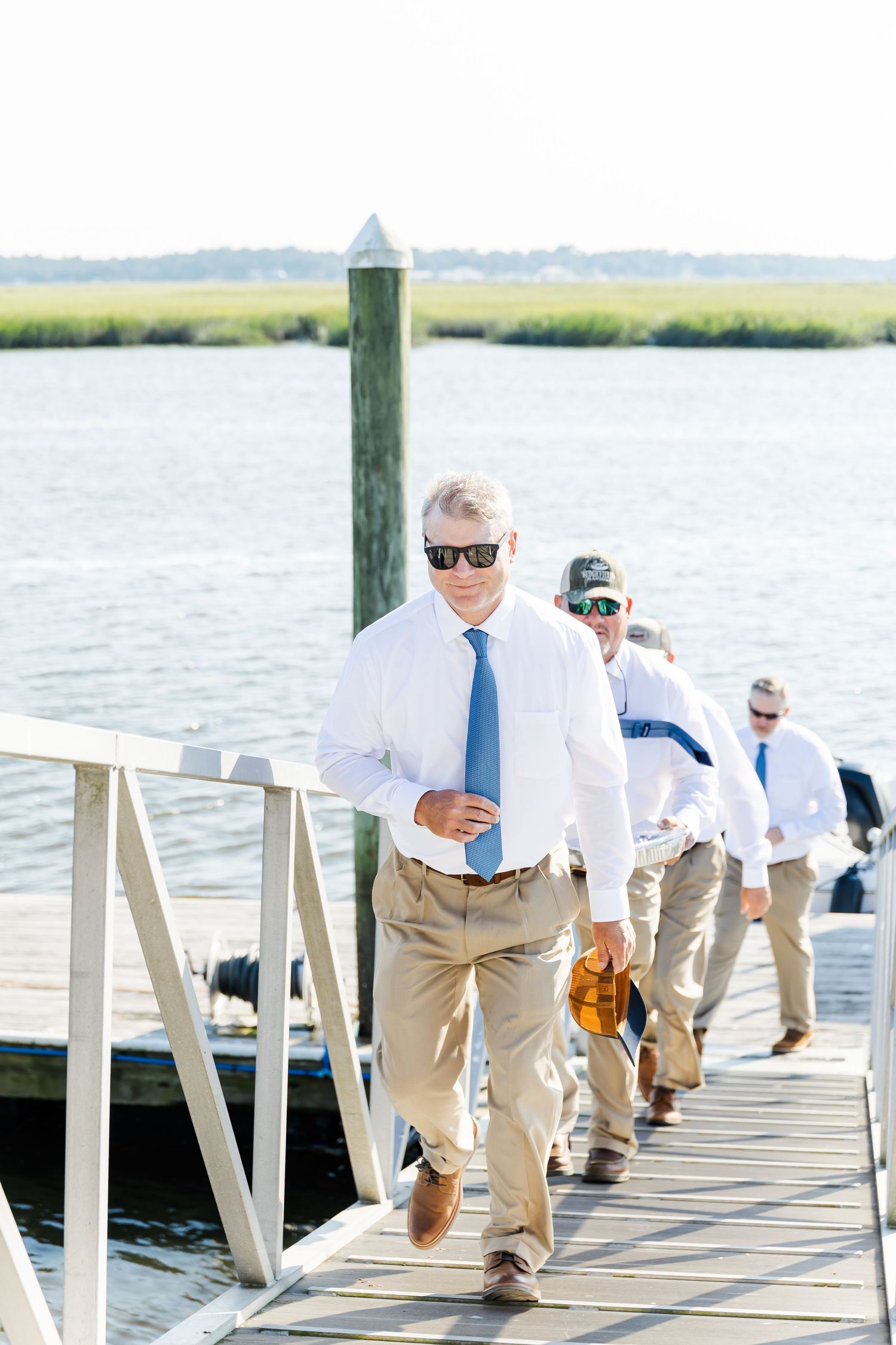 Groomsmen in khaki pants and sunglasses walk down a wooden dock by the water on a sunny day.