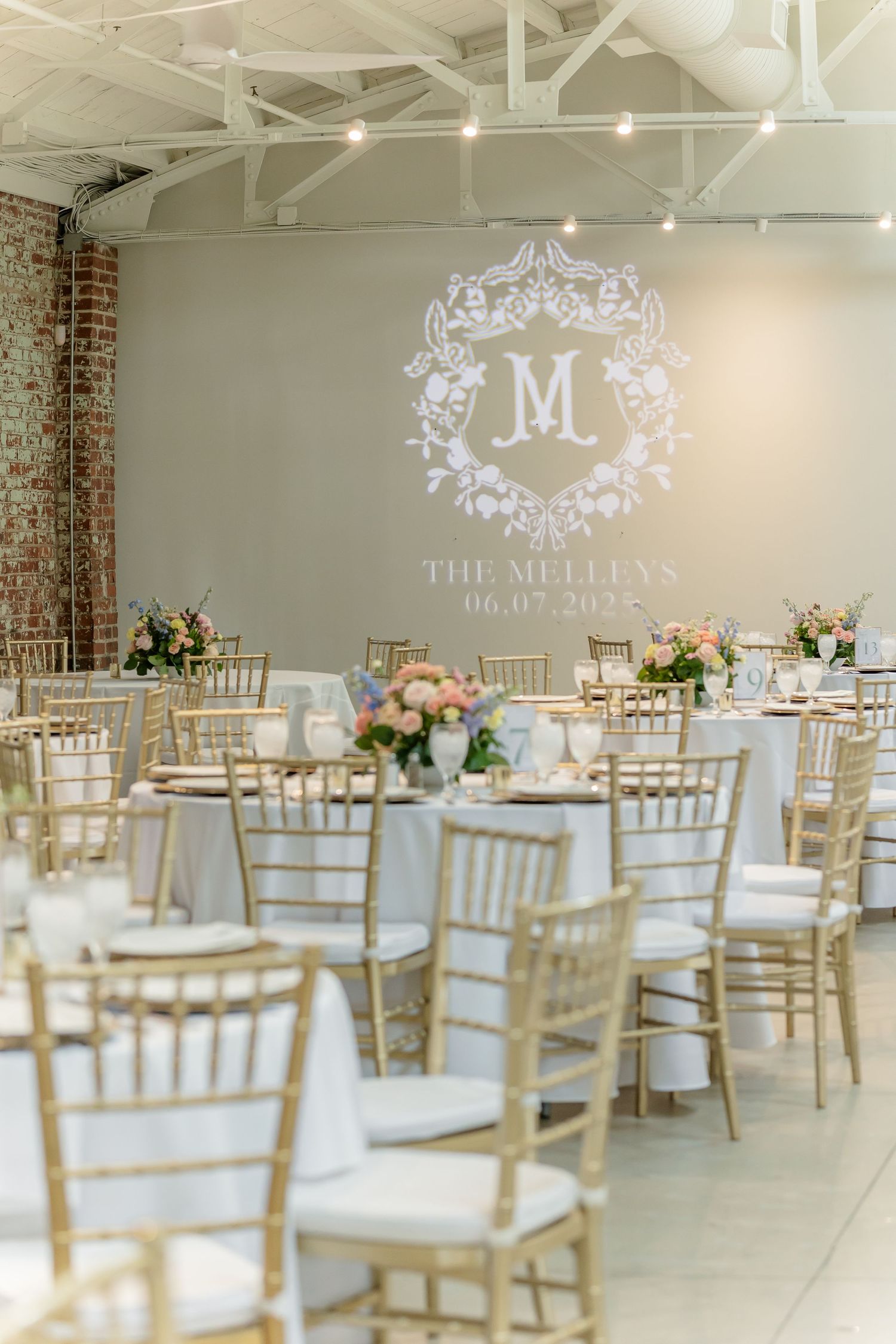 Elegant wedding reception venue with white walls, gold chiavari chairs, and ornate monogram projection on the wall.