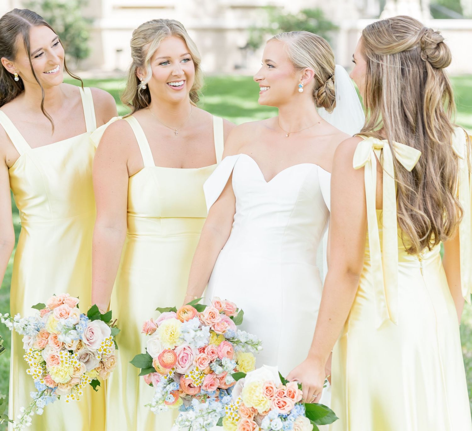 Bridesmaids in soft yellow dresses hold pastel bouquets while standing together outdoors for wedding photos.