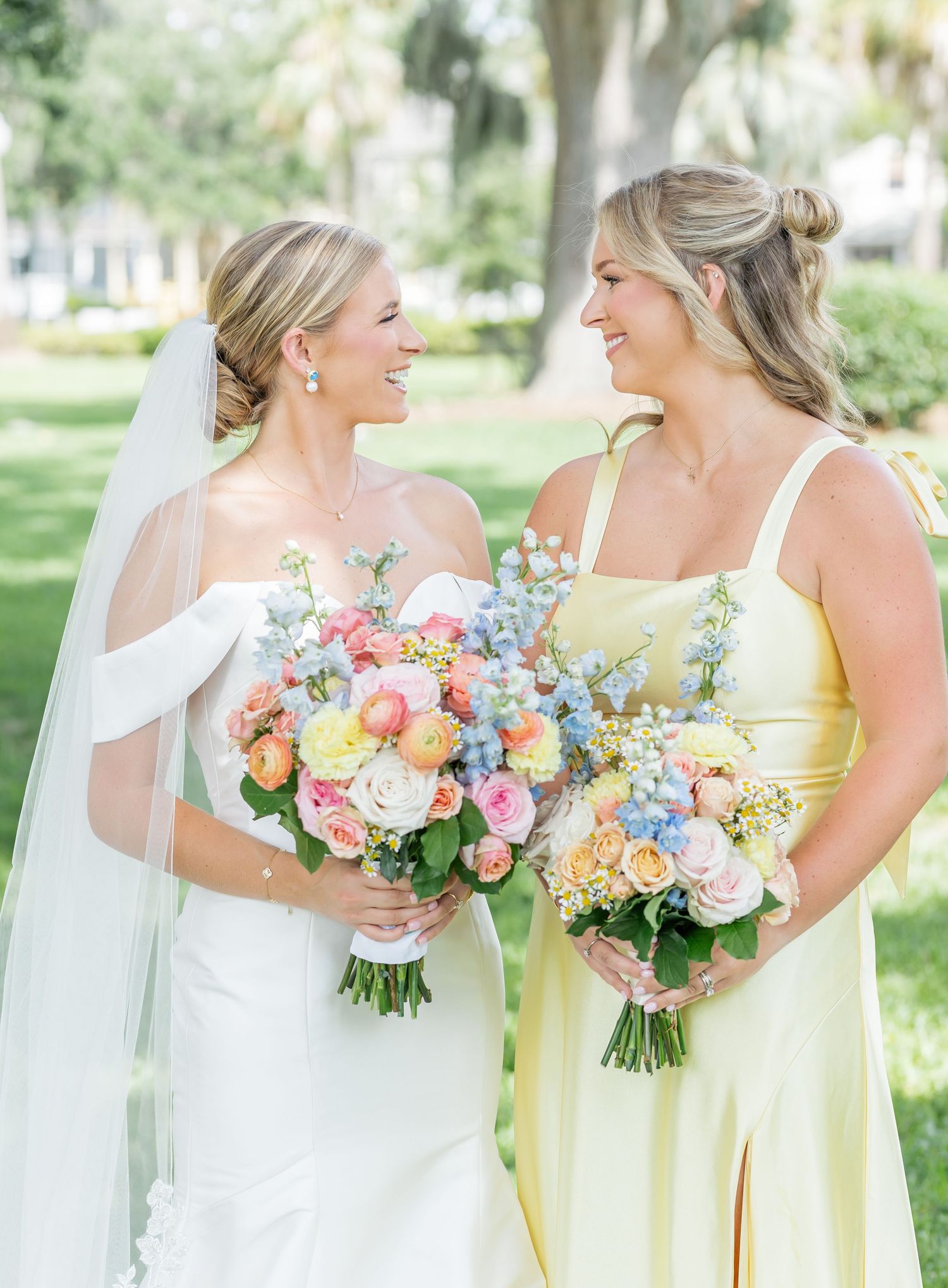 A bride in white and bridesmaid in yellow hold colorful spring bouquets while sharing joyful moments in a garden setting.
