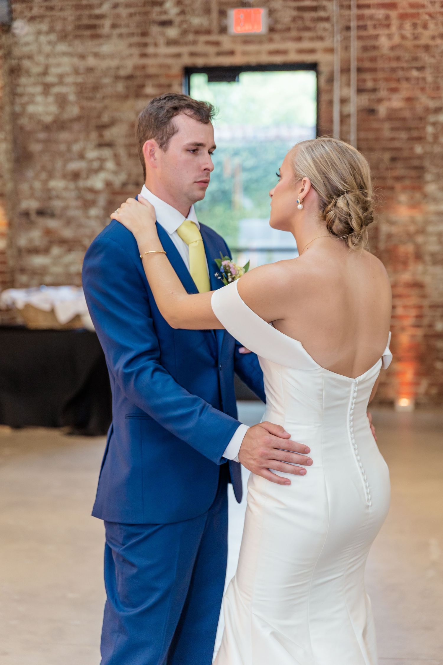 A couple shares their first dance in a rustic brick venue, with the bride in white and groom in navy blue.