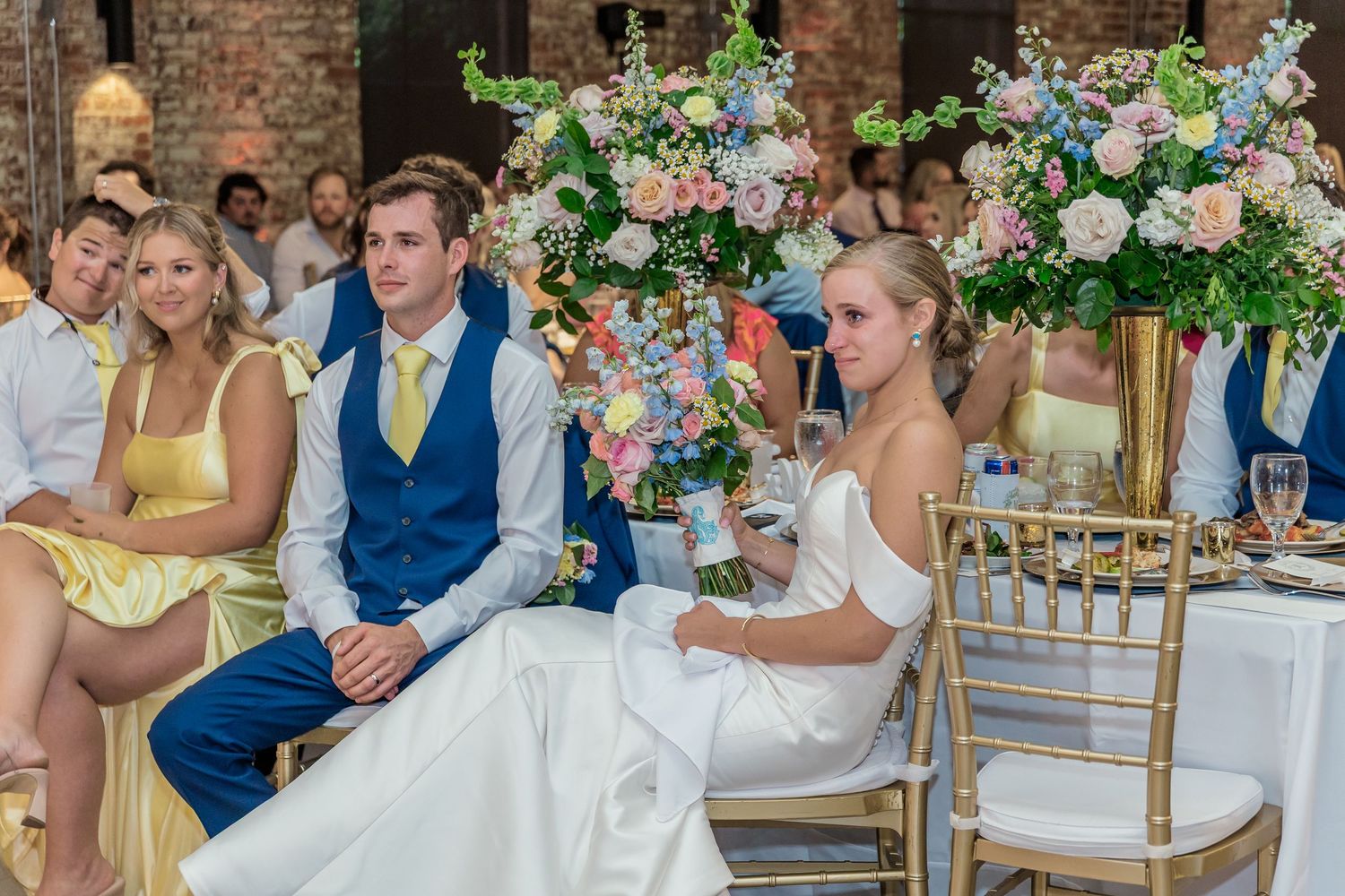 Wedding party members celebrate at a reception decorated with lush floral arrangements and exposed brick walls.
