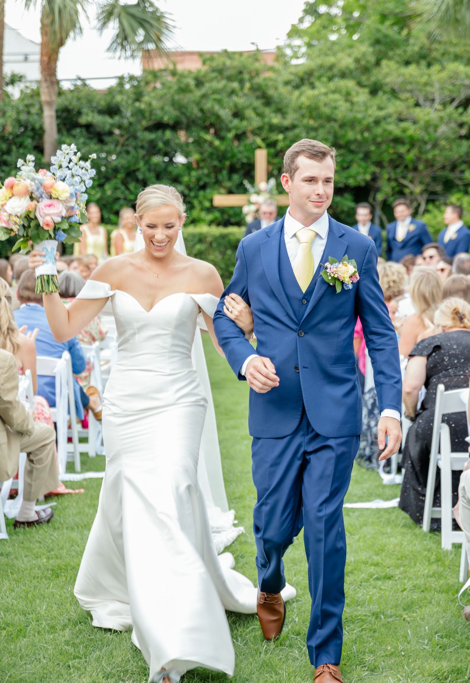 Wedding couple walks across a lush green lawn with guests seated on white chairs during an outdoor ceremony.