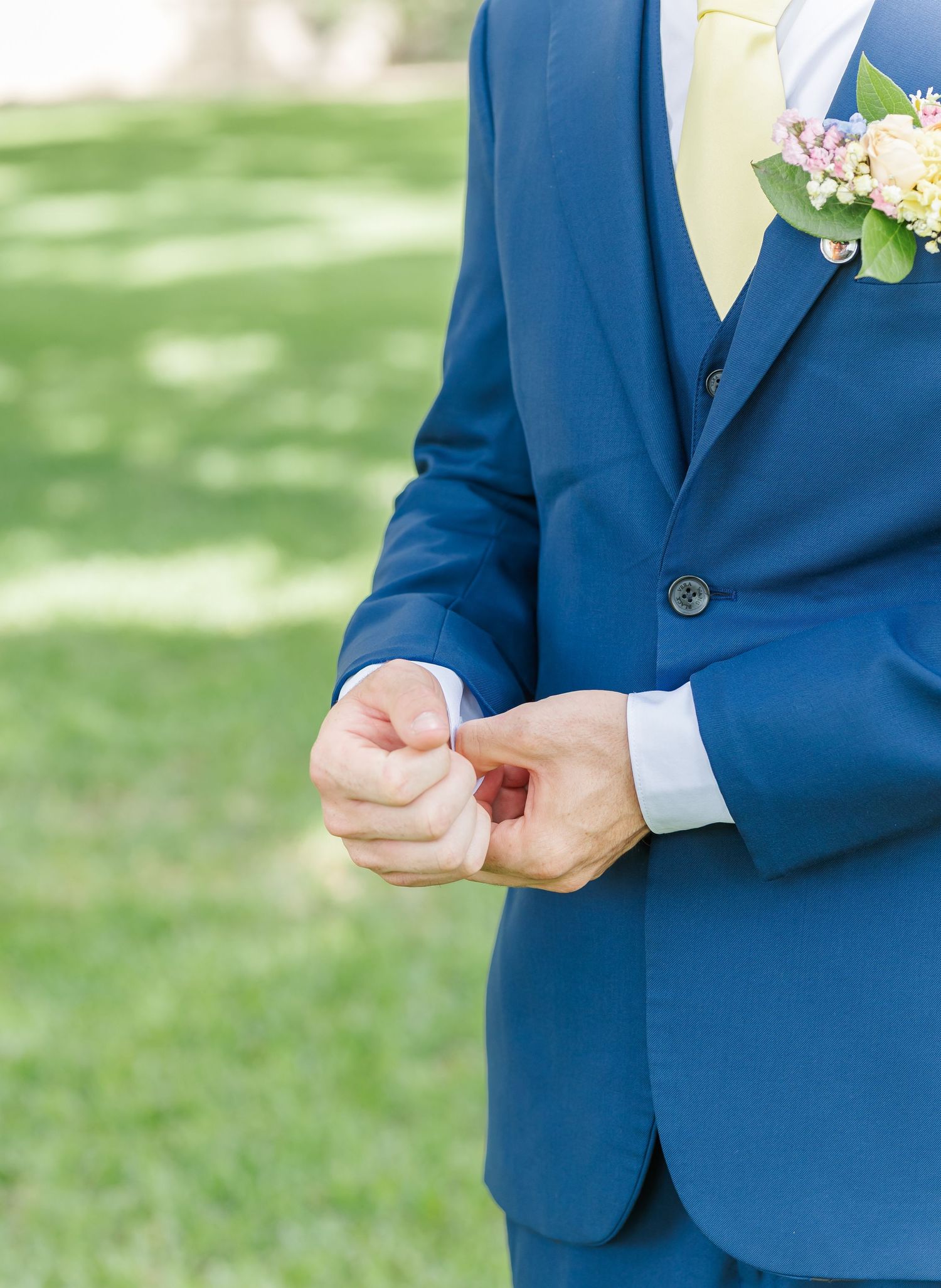 A groom in a navy blue suit adjusts his boutonniere while standing outdoors on a grassy lawn.